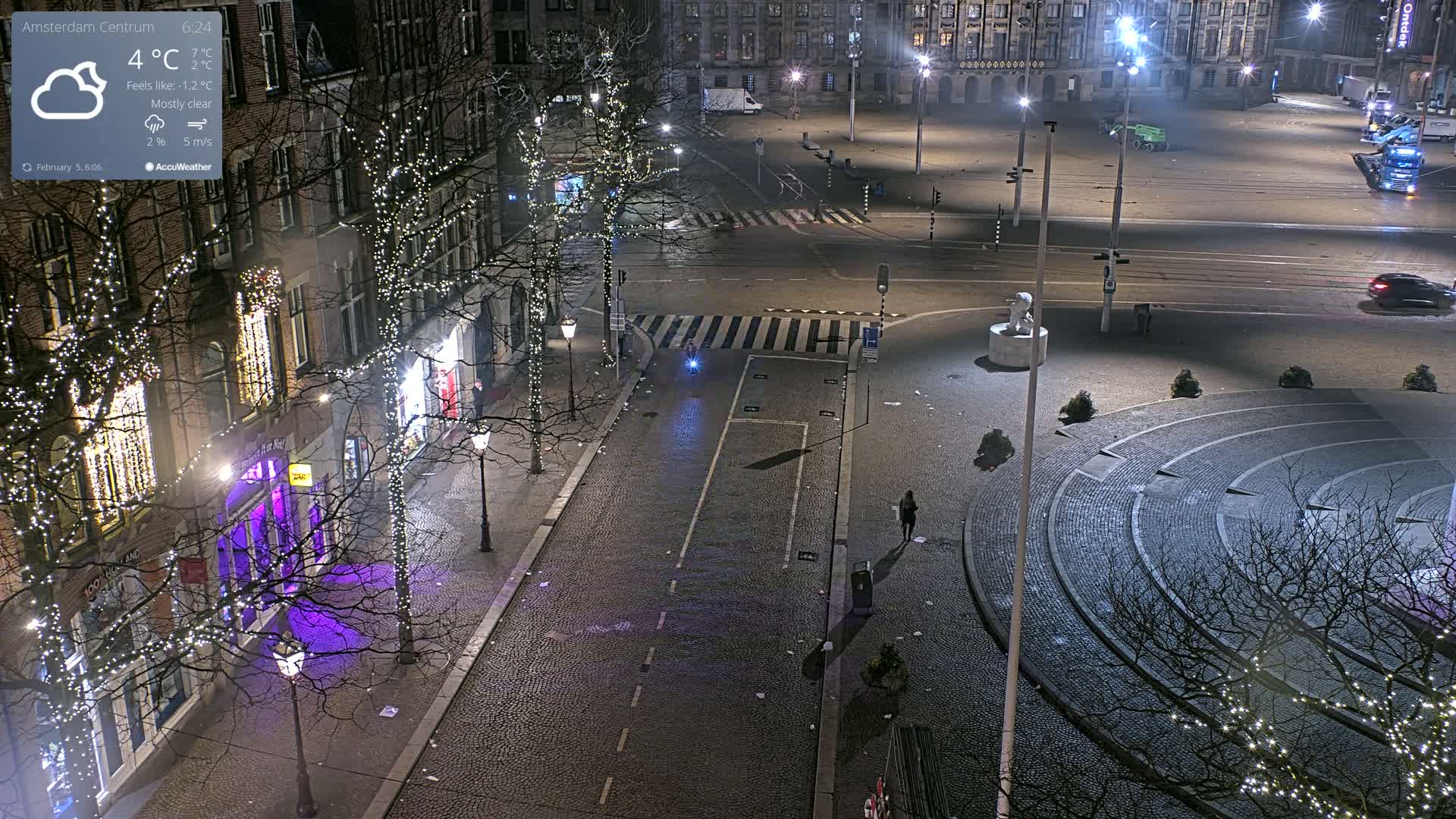 A mostly clear, cold night scene of a city square with sparsely populated streets, buildings adorned with Christmas lights, and a circular paved area.