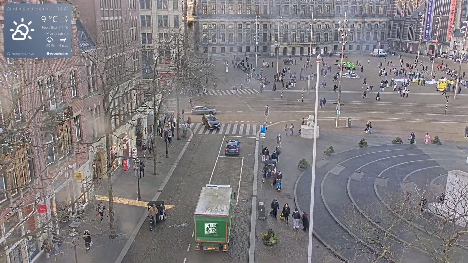 A mostly sunny city square with a few pedestrians and several cars and a truck on the street, viewed from an elevated position.