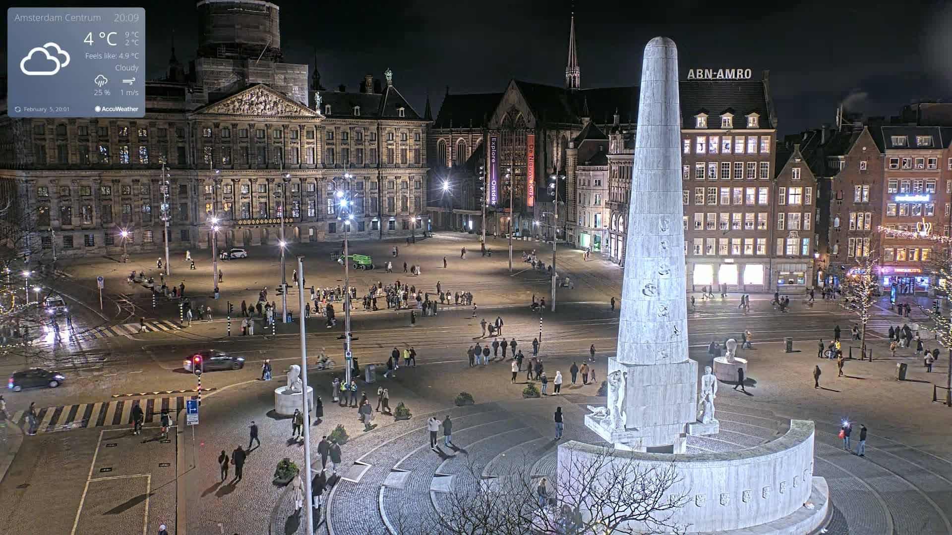 A nighttime, cloudy view of a large city square with a monument at its center, surrounded by buildings and people.