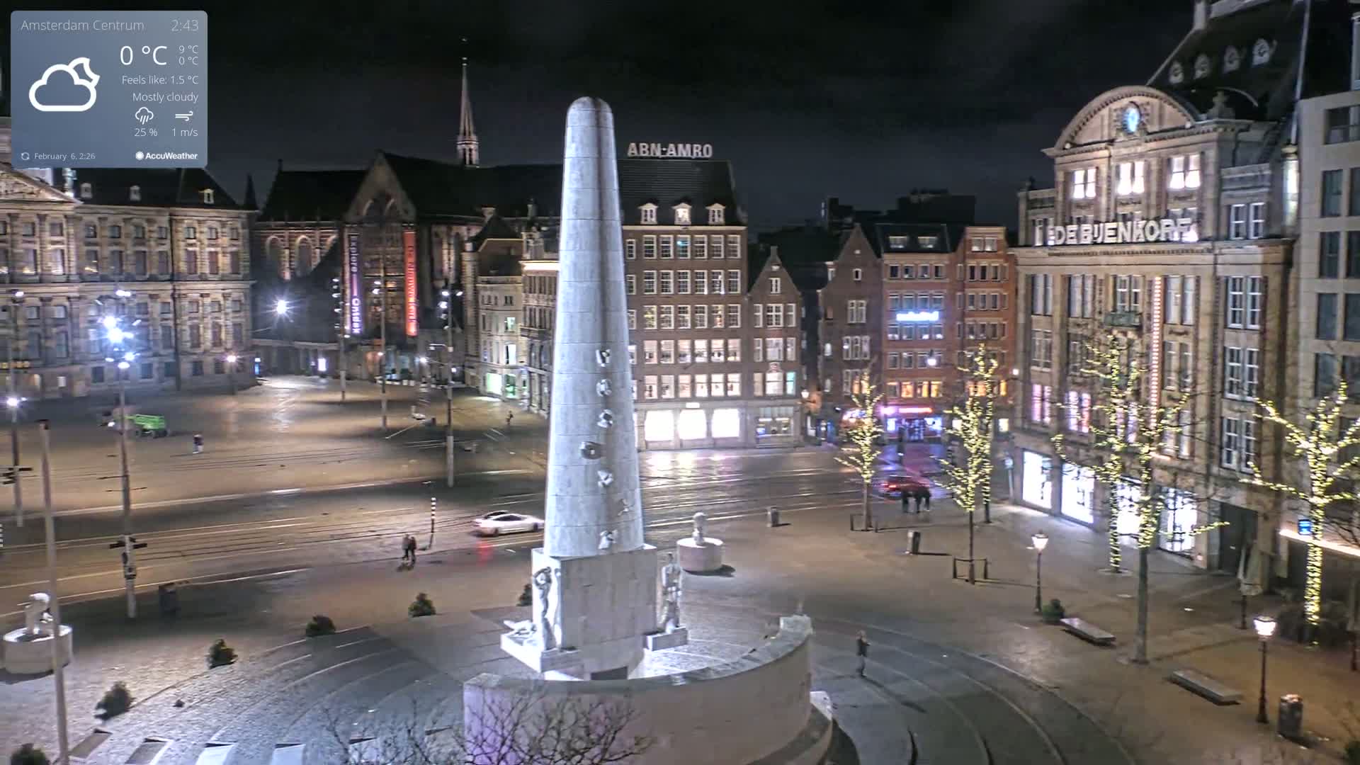 A mostly cloudy night scene shows a tall monument in a city square surrounded by buildings with illuminated windows and streetlights, some trees with string lights, and a few pedestrians.