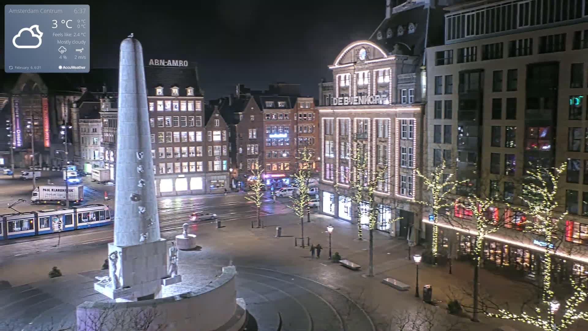 A mostly cloudy night scene shows a city square with a tall monument at its center, surrounded by illuminated buildings and trees decorated with lights, a tram, and cars.