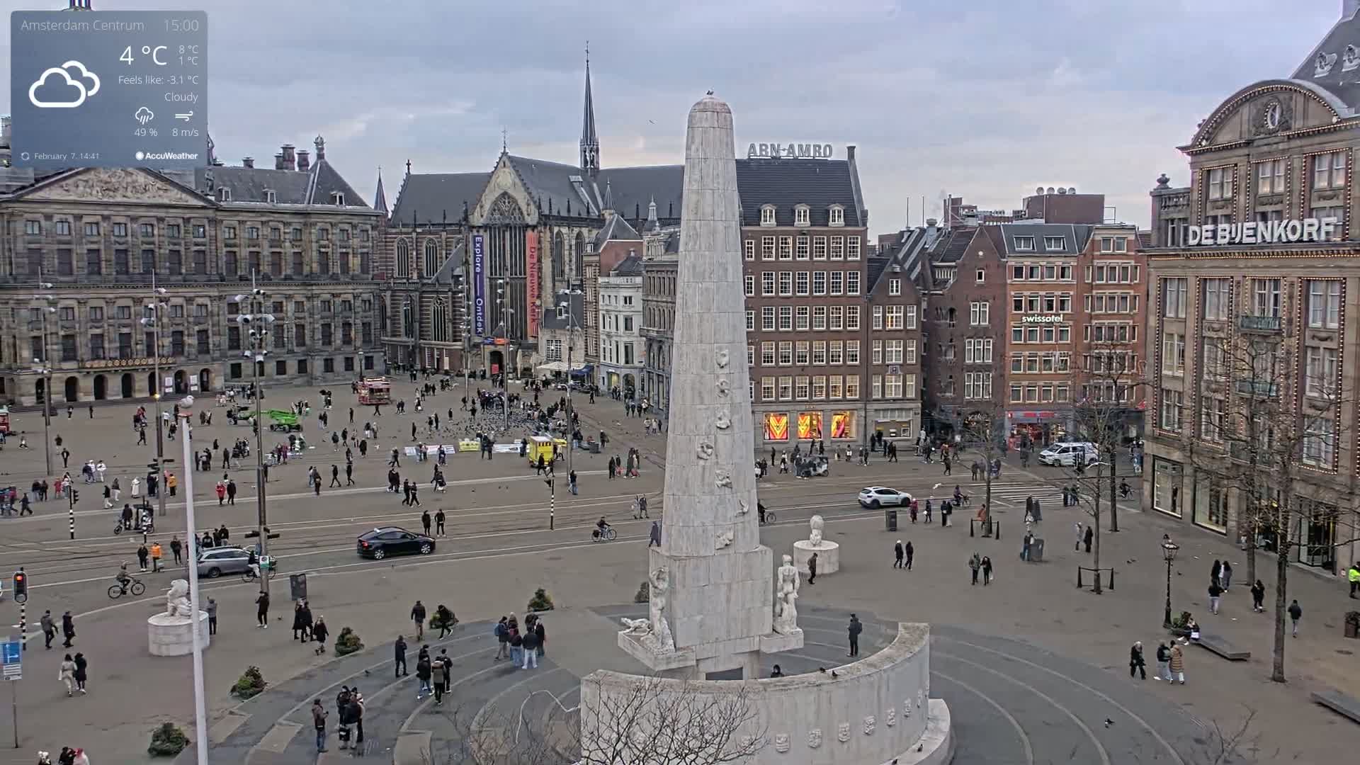 A cloudy, chilly day in a large city square with a tall monument at its center, surrounded by historic buildings and pedestrians.