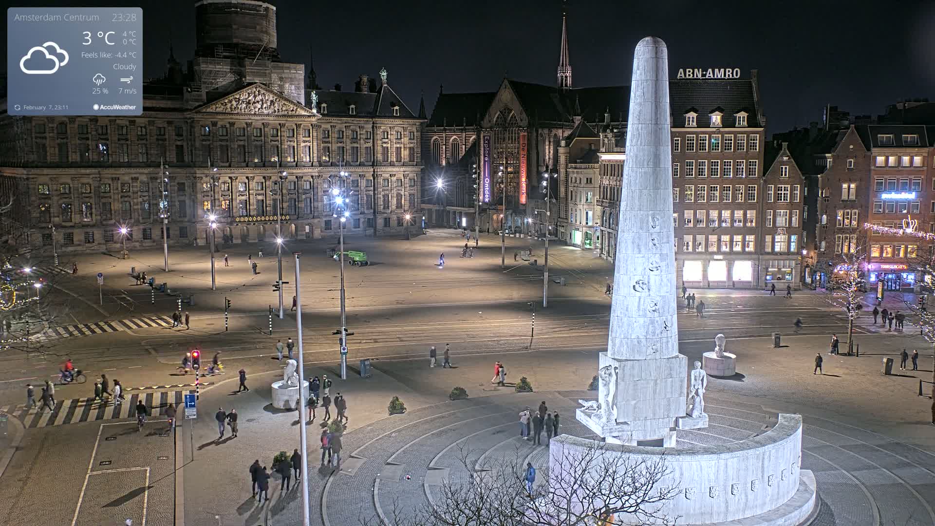 A nighttime, cloudy view of a large city square with a tall monument at its center, surrounded by buildings, and sparsely populated with pedestrians and cyclists.