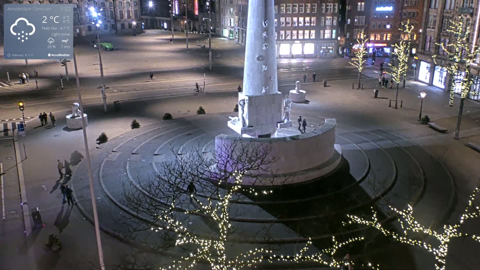 A nighttime view of a city square with a tall monument at its center, under light snow with some pedestrians visible.