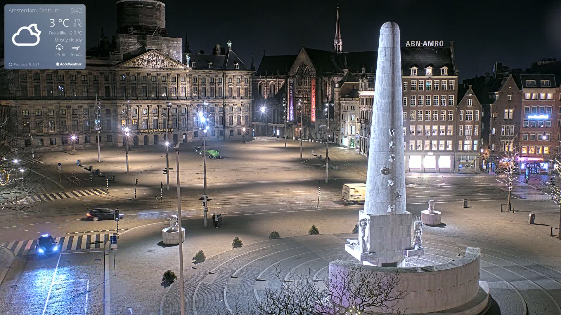 A mostly cloudy night scene shows a large, empty city square with a tall monument at its center, surrounded by historic buildings, illuminated by streetlights.