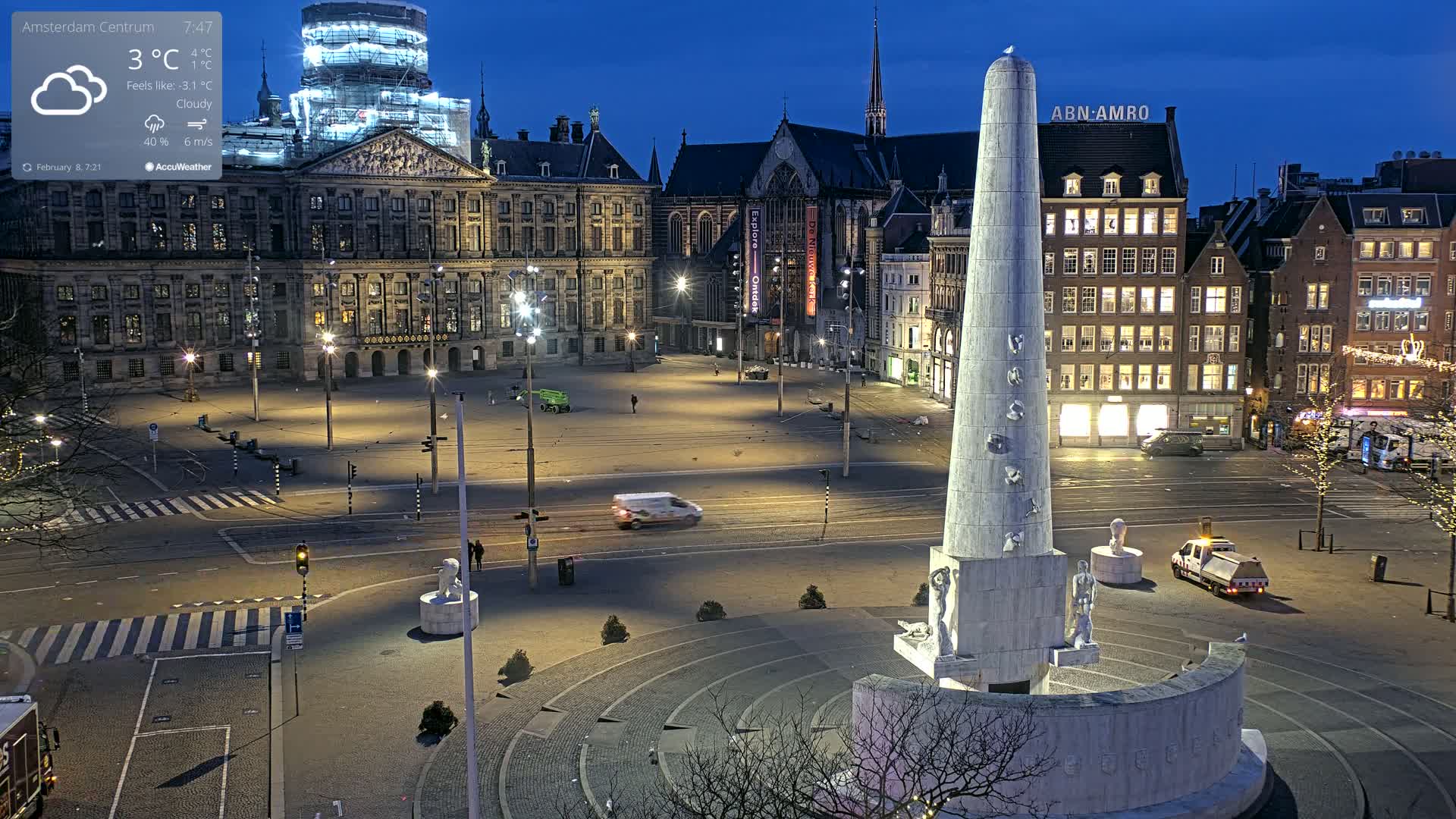 A mostly empty city square at night under cloudy skies, featuring a large monument at its center, several buildings, and a few vehicles.