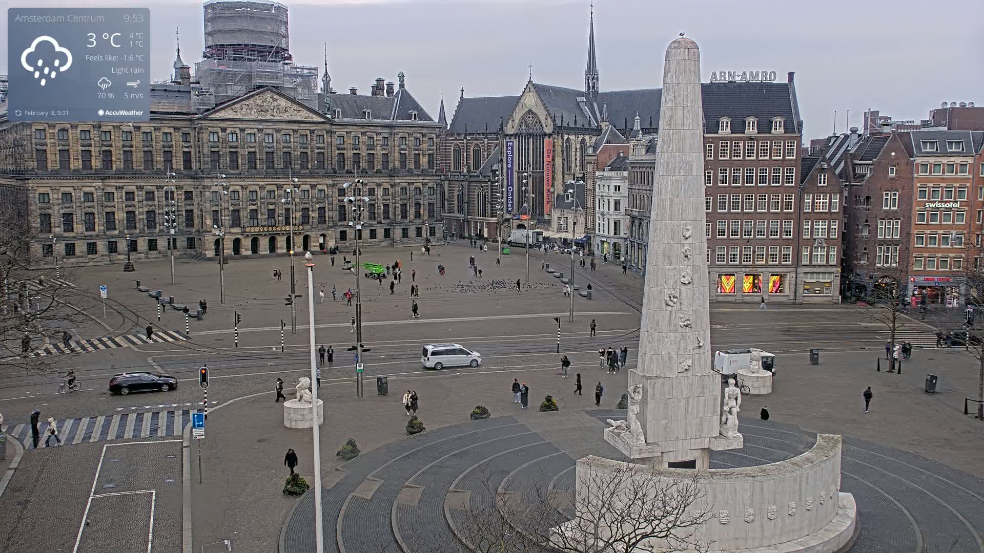 A city square with a tall monument at its center, surrounded by buildings under a light rain.