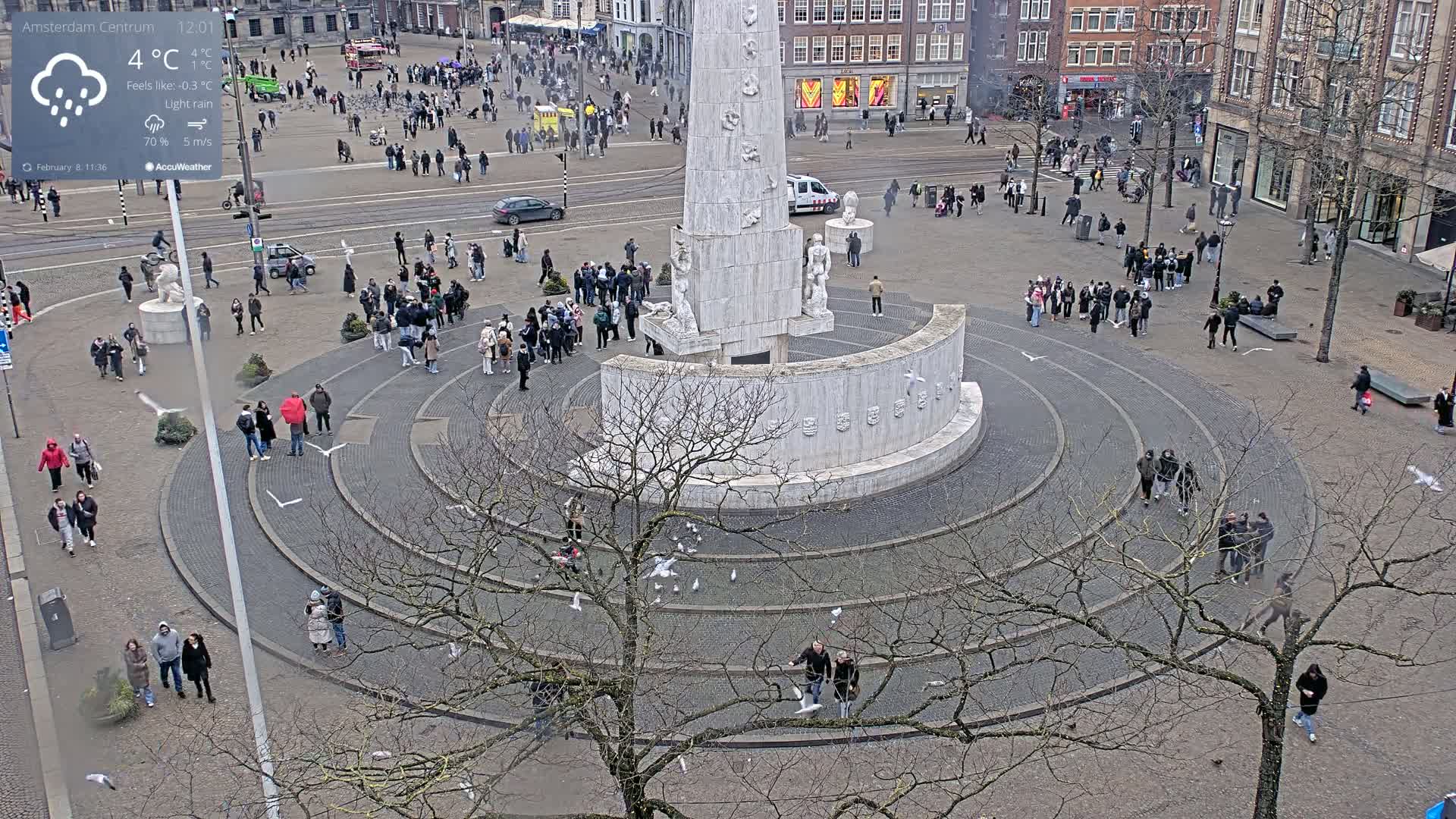 A large, circular plaza surrounds a tall, white monument, with many people scattered throughout the area under light rain.