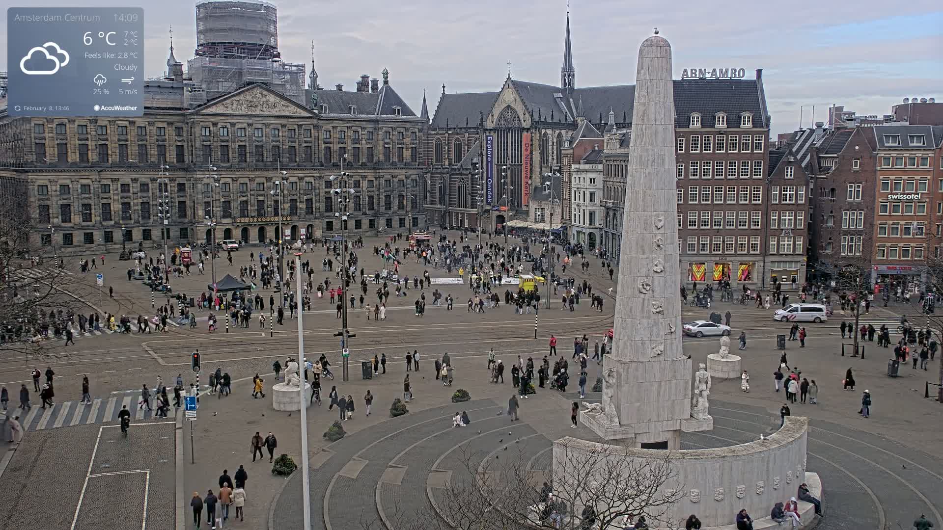 A cloudy day in a large European city square, centered around a tall, white monument, with many people walking around and buildings surrounding the square.