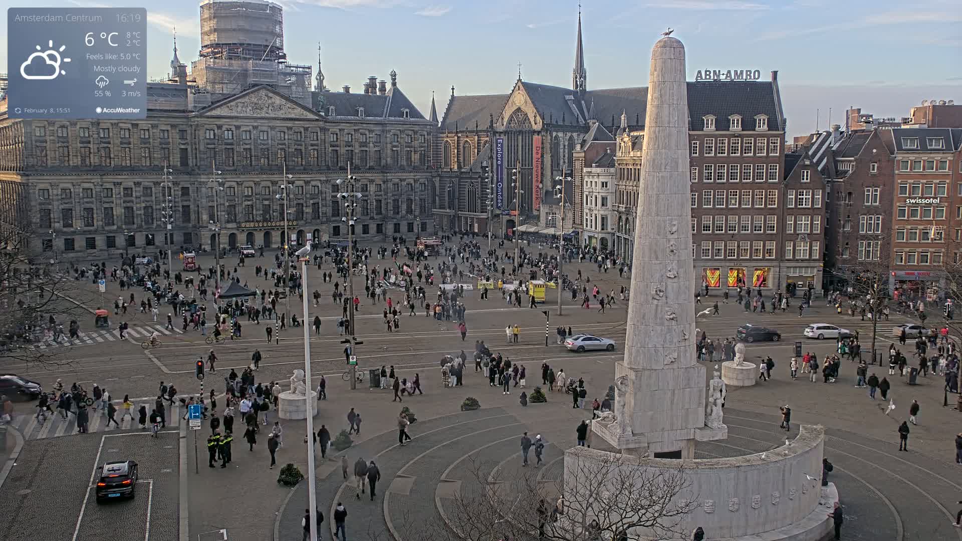A mostly cloudy day in a large city square, centered on a tall, light-colored monument surrounded by many people, buildings, and cars.