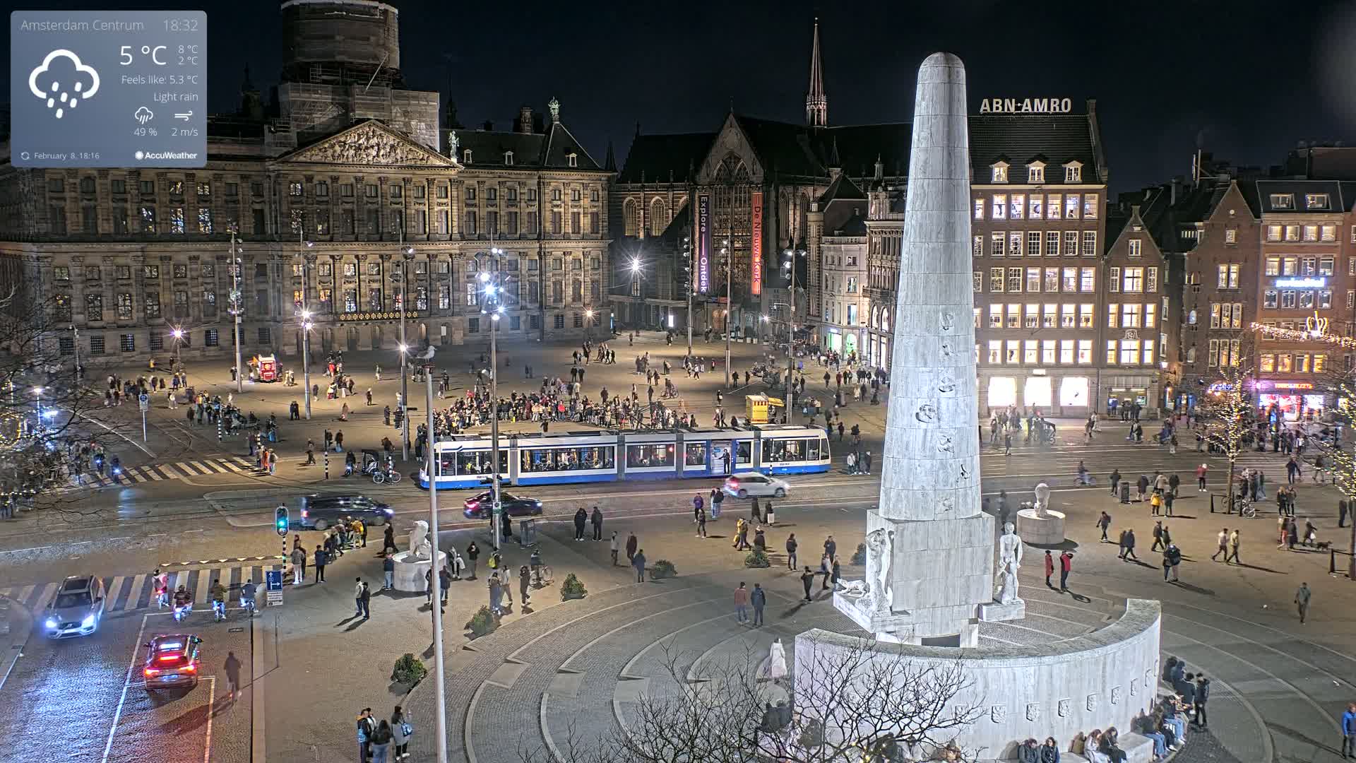 A nighttime, lightly raining city square scene shows a tram passing near a large monument, with people walking around the square and buildings lit up around the perimeter.