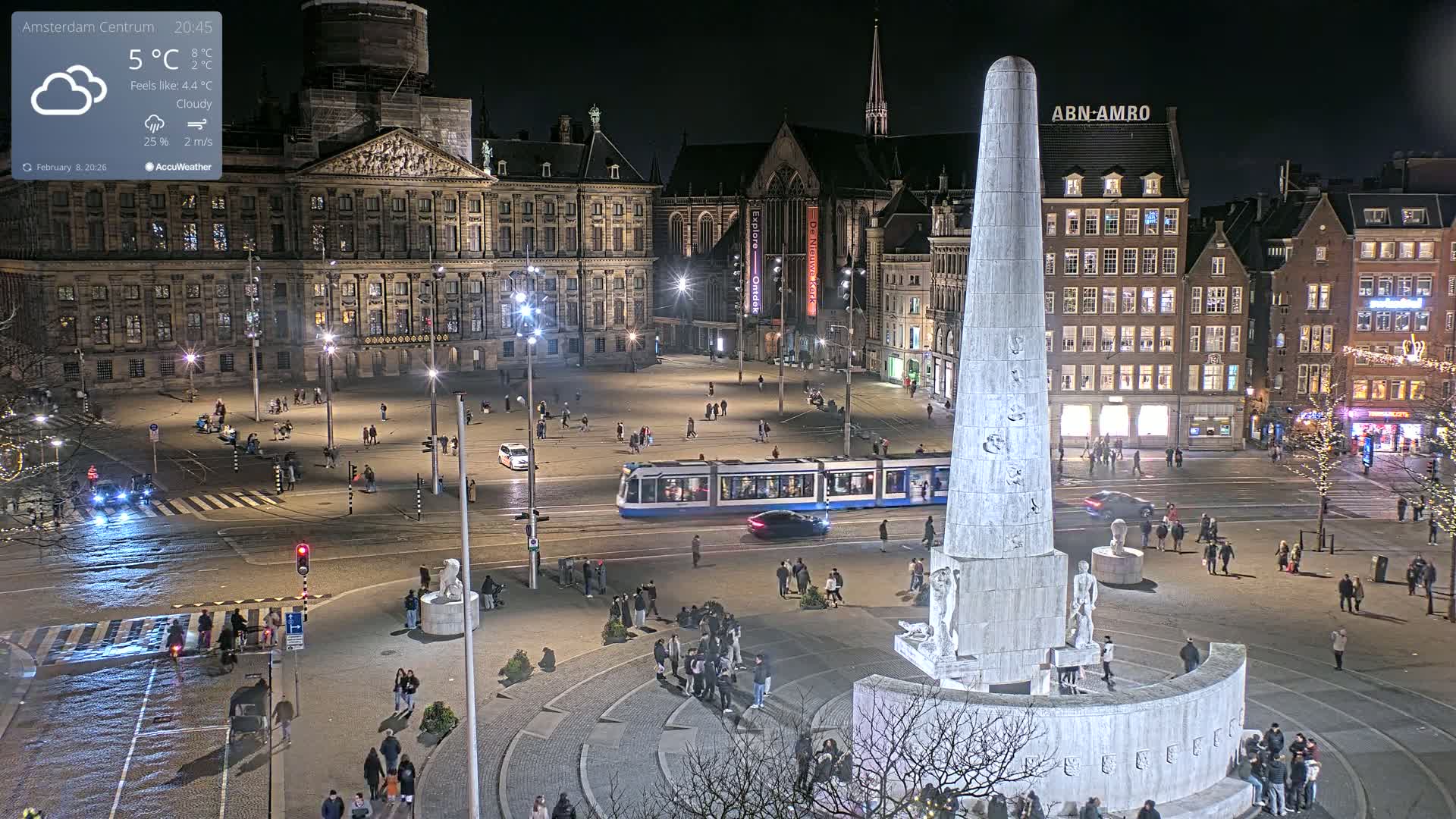 A nighttime, cloudy view of a city square with a tall monument at its center, surrounded by buildings, a tram, and pedestrians.
