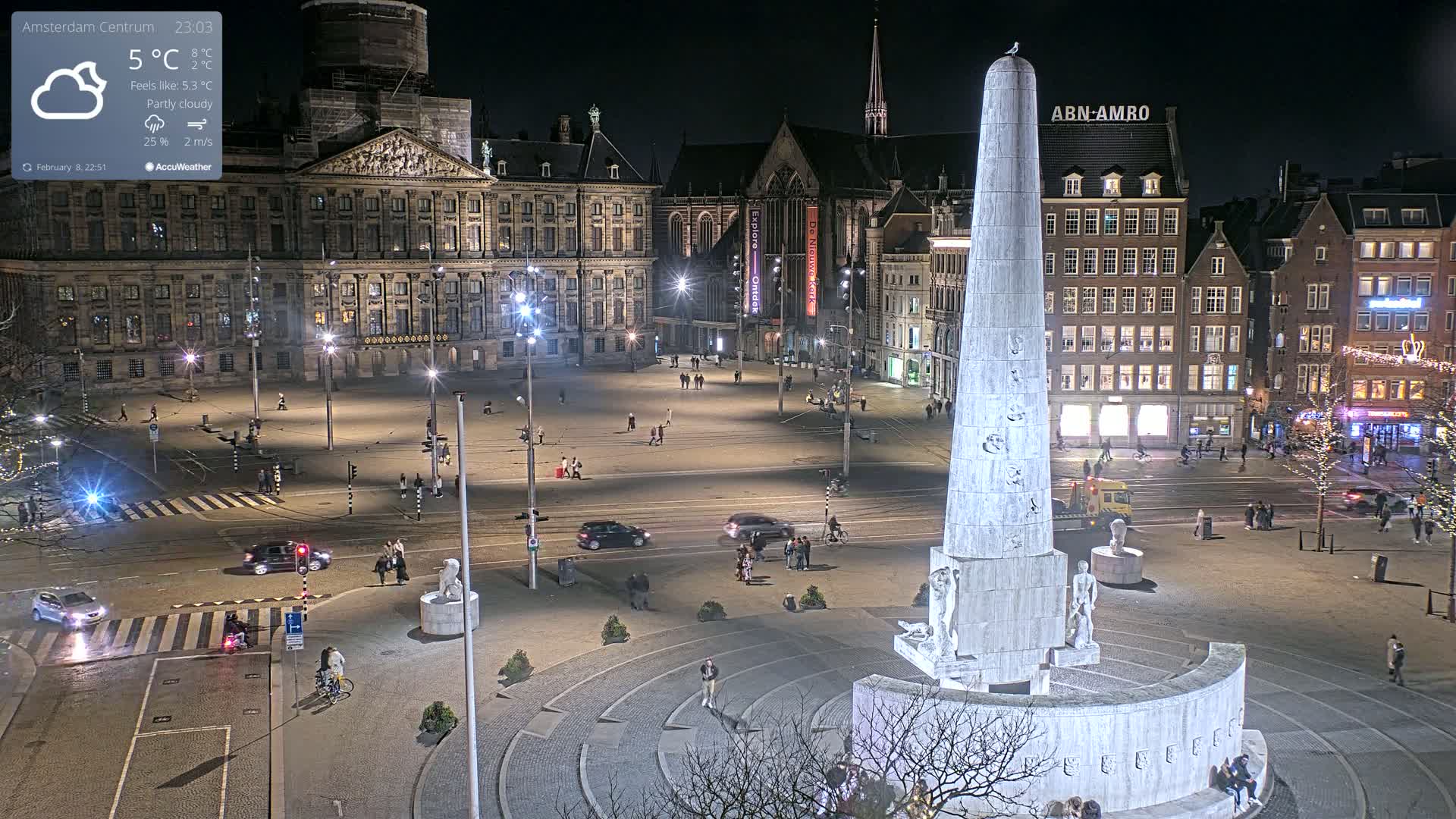 A nighttime, partly cloudy view of a city square with a tall monument at its center, surrounded by buildings and sparse pedestrian traffic.
