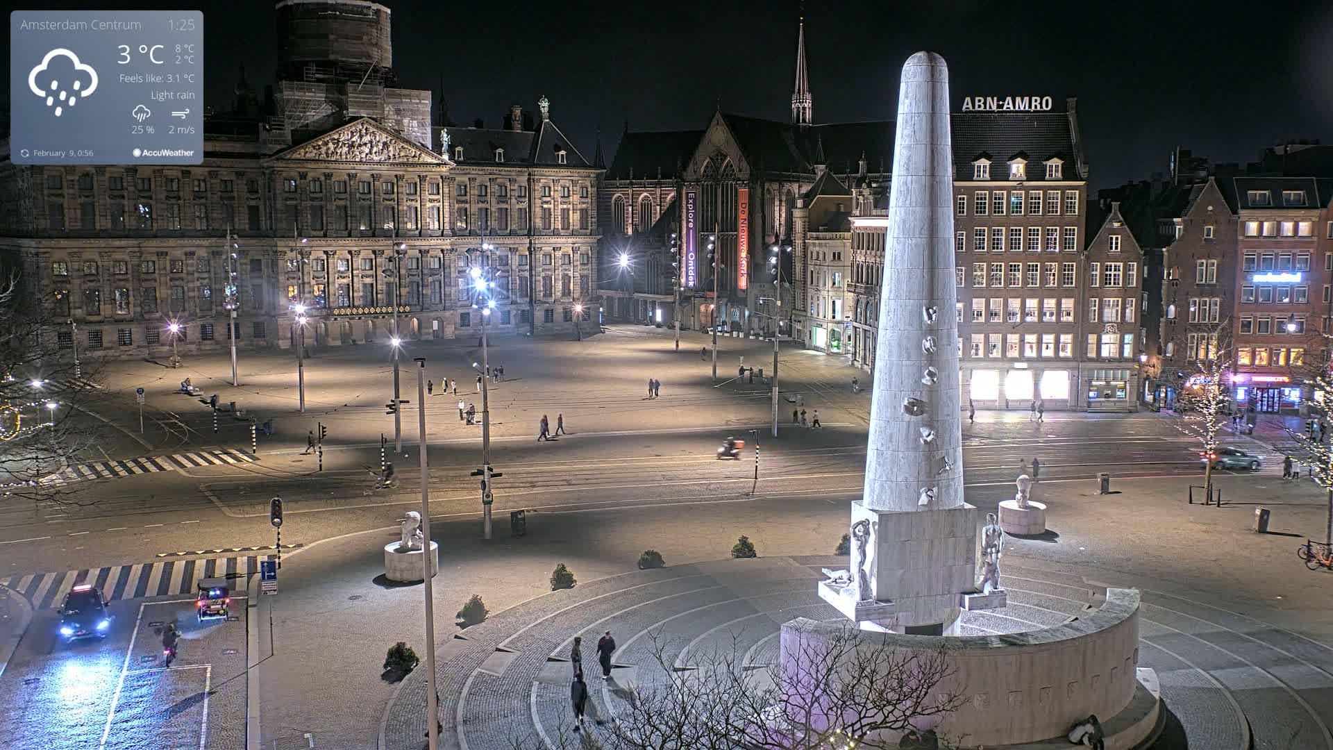 A nighttime view of a large, sparsely populated plaza with a tall monument at its center, under conditions of light rain.