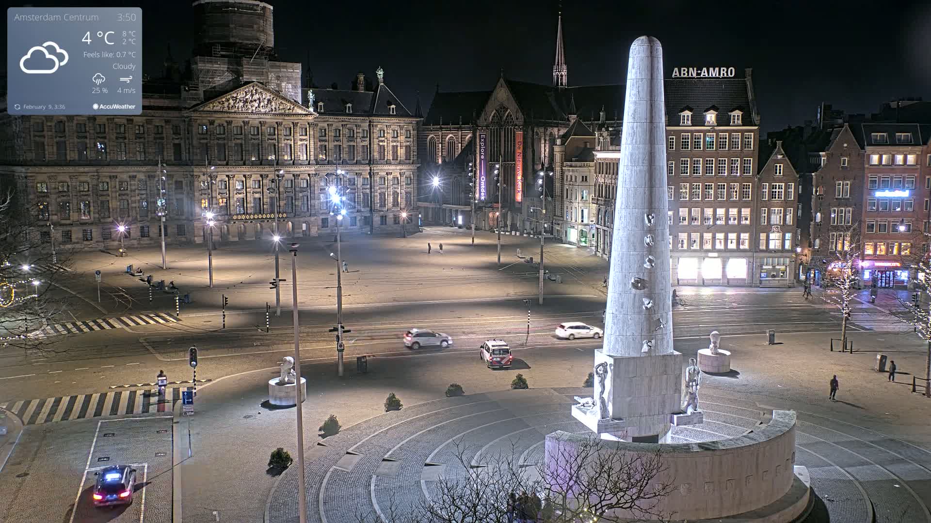 A nighttime, cloudy view of a large, sparsely populated city square with a tall monument at its center, surrounded by buildings, including a large palace.