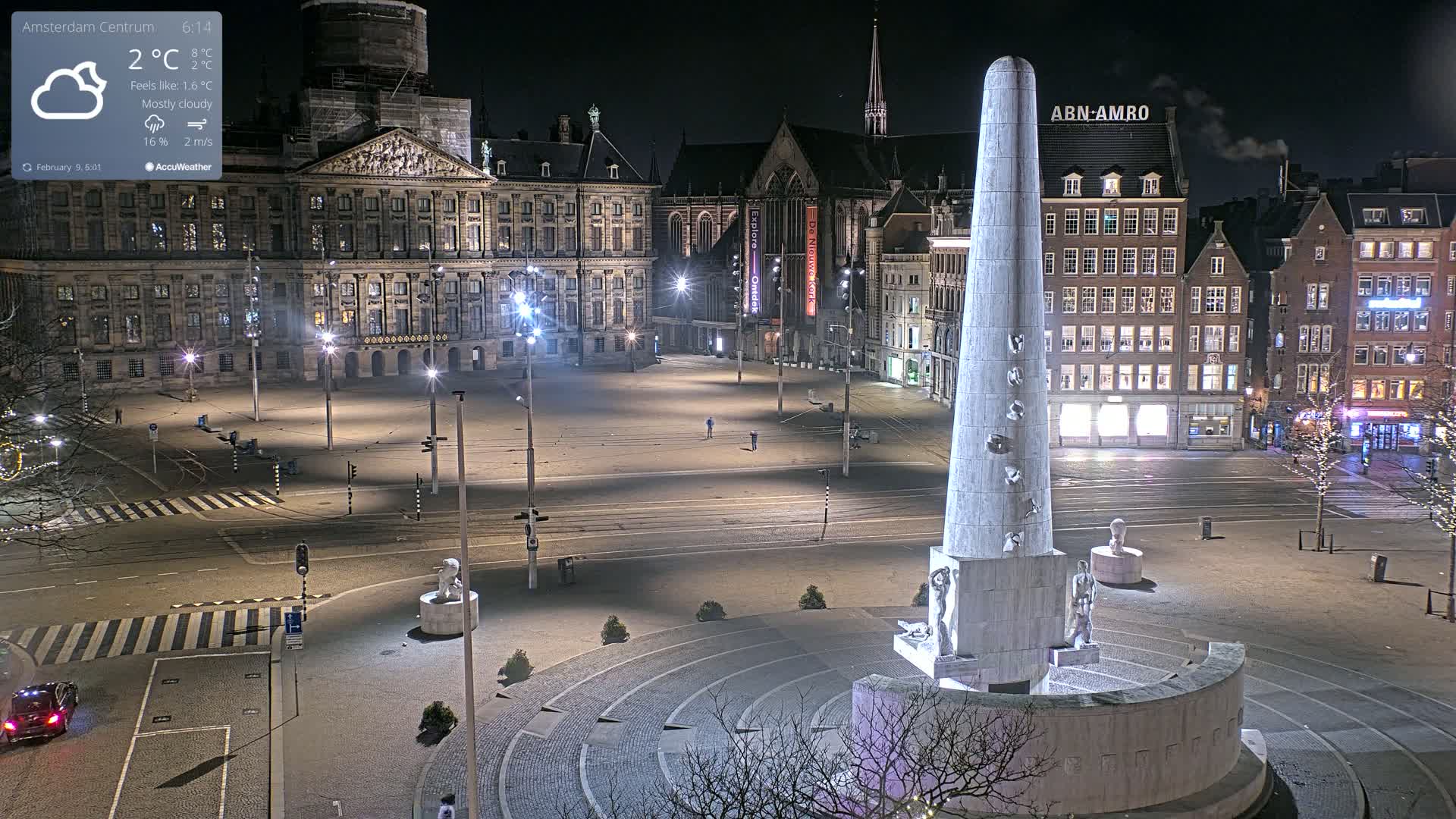 A mostly cloudy night scene shows a large, empty city square with a tall monument in the center, surrounded by buildings illuminated by streetlights.