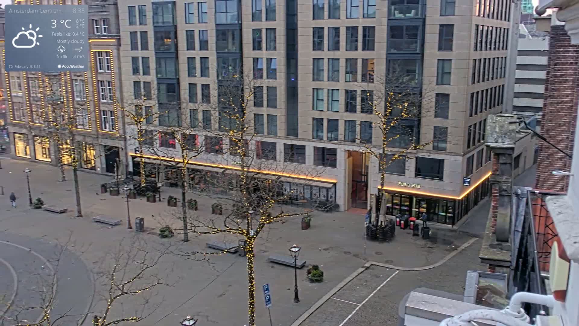 A mostly cloudy, cool day in a city square with bare trees decorated with string lights, and several buildings visible.