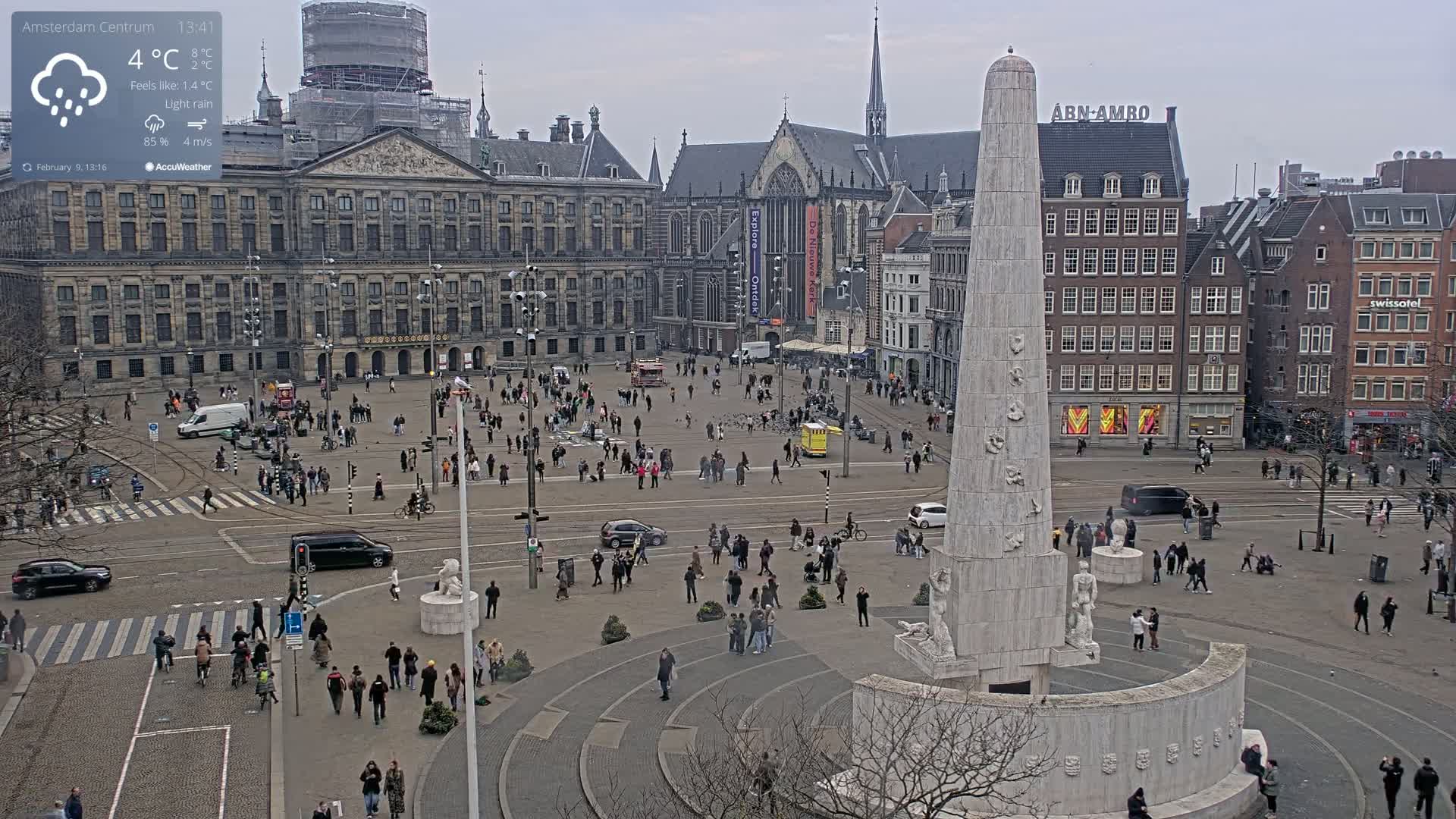 A wide shot of a city square with a tall monument in the foreground under light rain, surrounded by pedestrians and buildings, including a large palace.