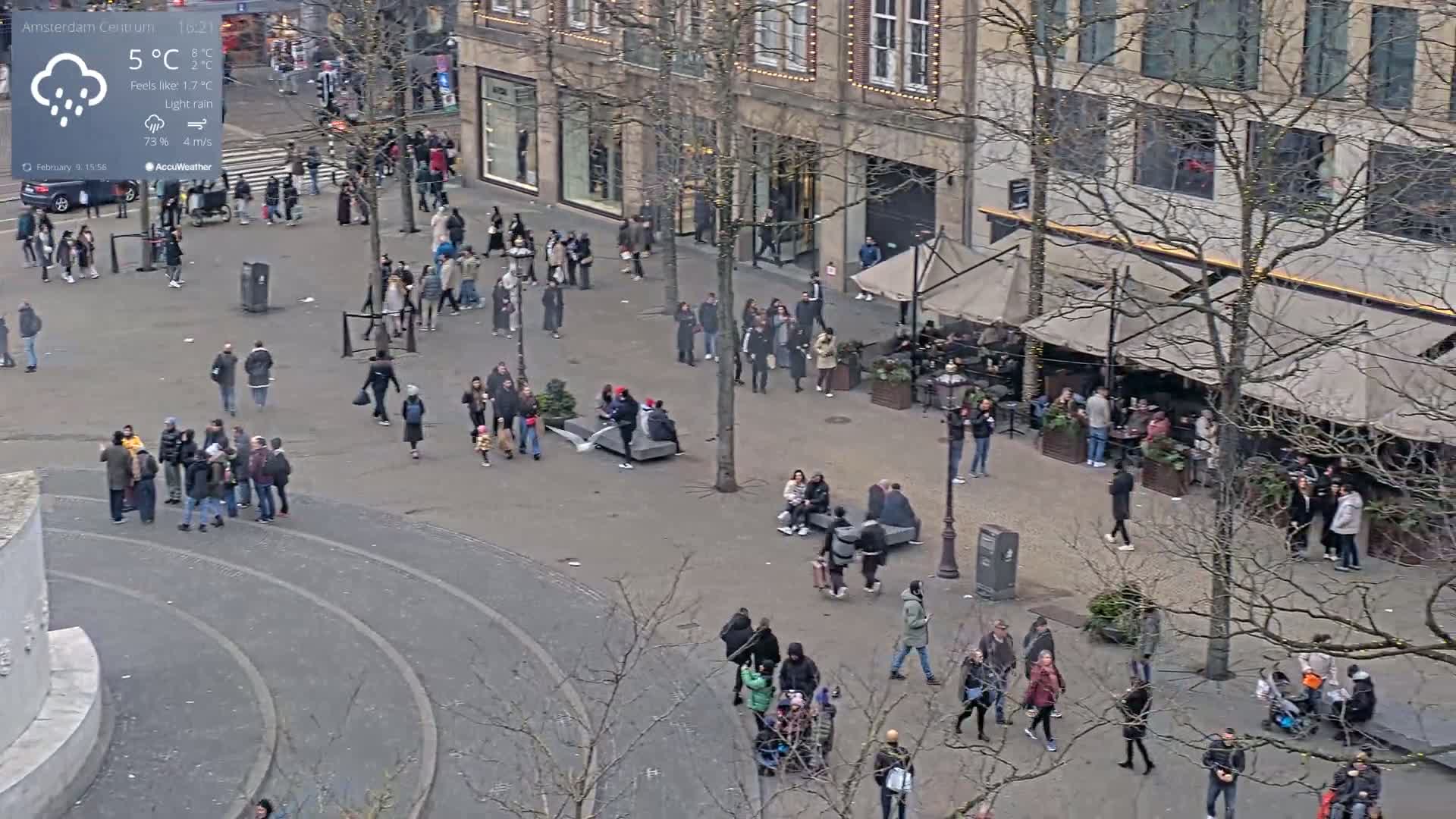A city square with many people walking around under light rain, near buildings with outdoor seating areas.