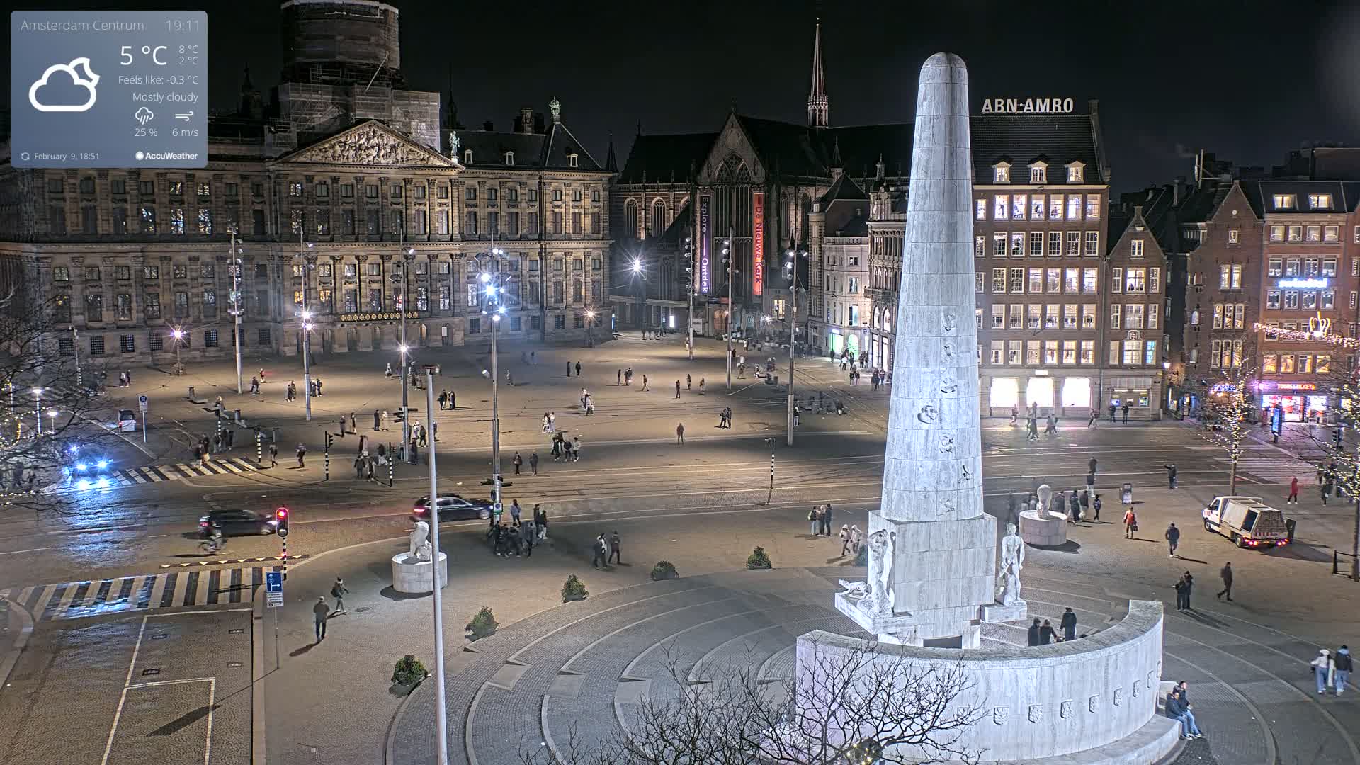 A nighttime, mostly cloudy view of a large city square with a tall monument at its center, surrounded by buildings and sparsely populated by pedestrians.