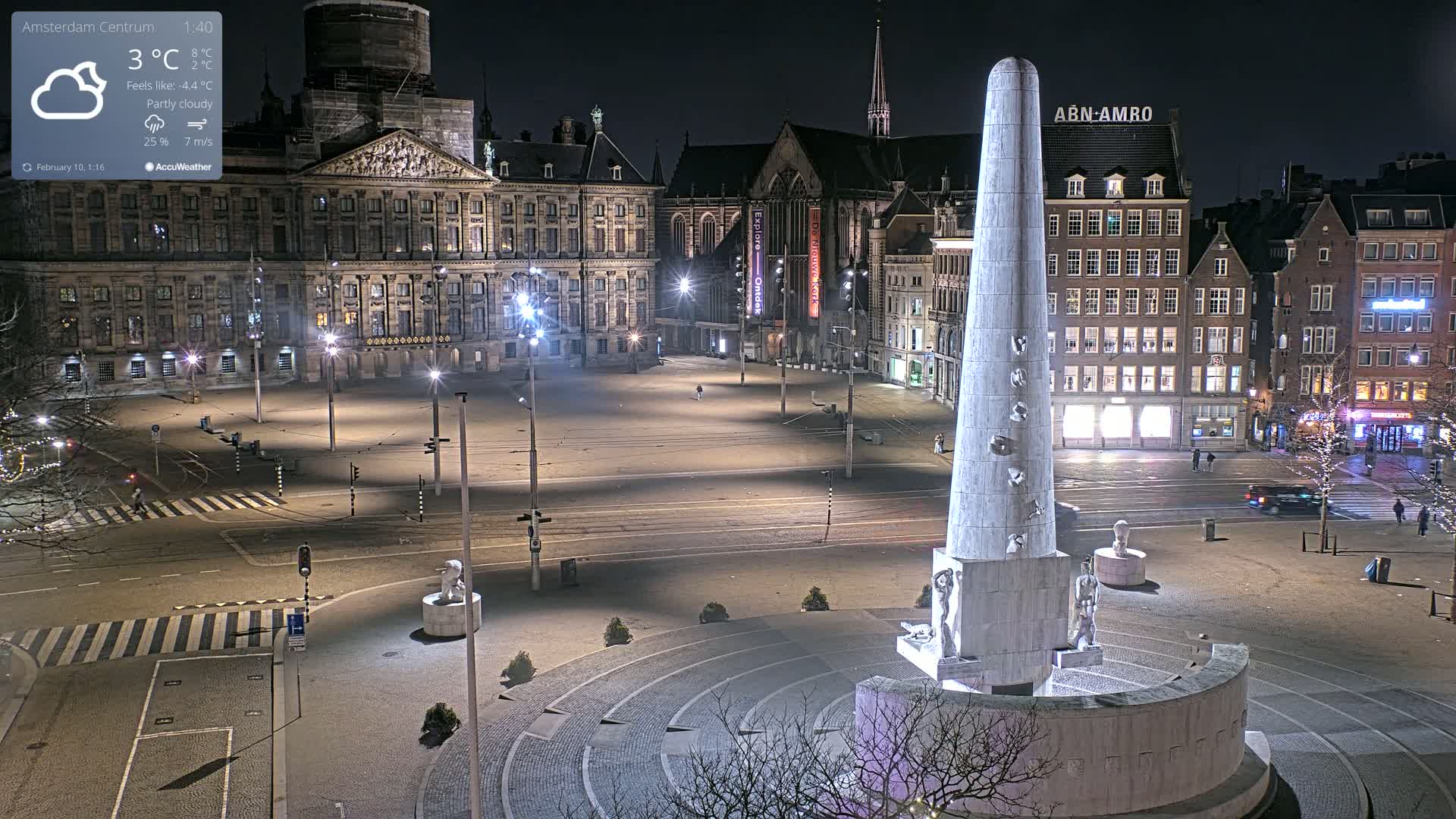 A nighttime, partly cloudy view of a large city square with a tall monument in the center, surrounded by historical buildings and illuminated streets.