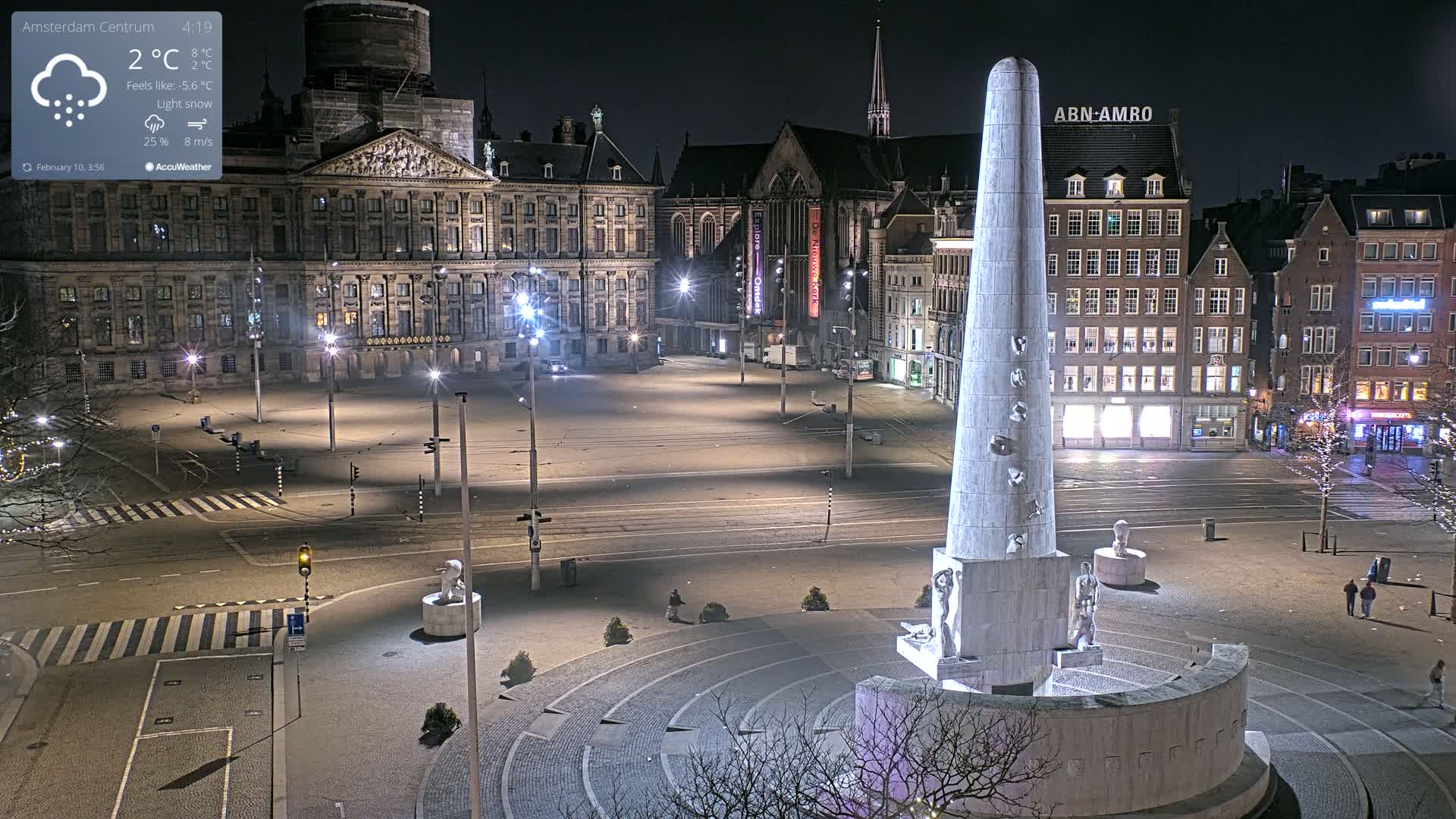 A nighttime, lightly snowing city square with a tall monument at its center, surrounded by historical buildings and sparsely populated with people.