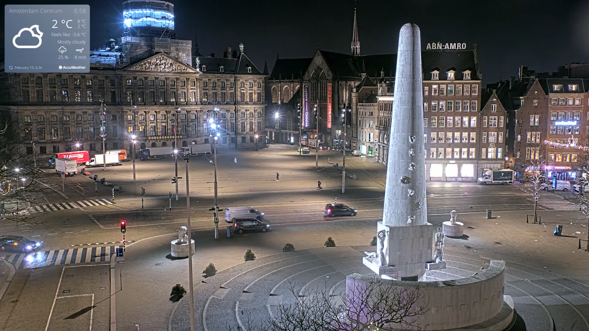 A mostly cloudy night scene shows a large city square with a tall monument at its center, surrounded by buildings, some vehicles, and a few pedestrians.