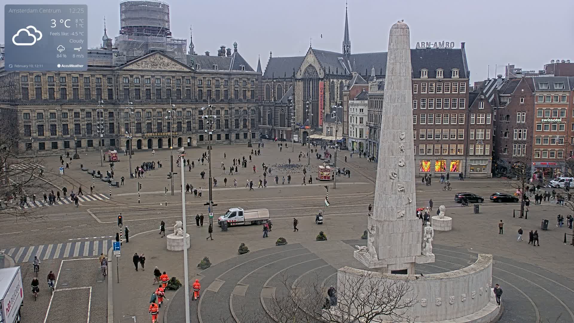 A cloudy, cold city square with a large monument in the center, surrounded by historical buildings and people walking.