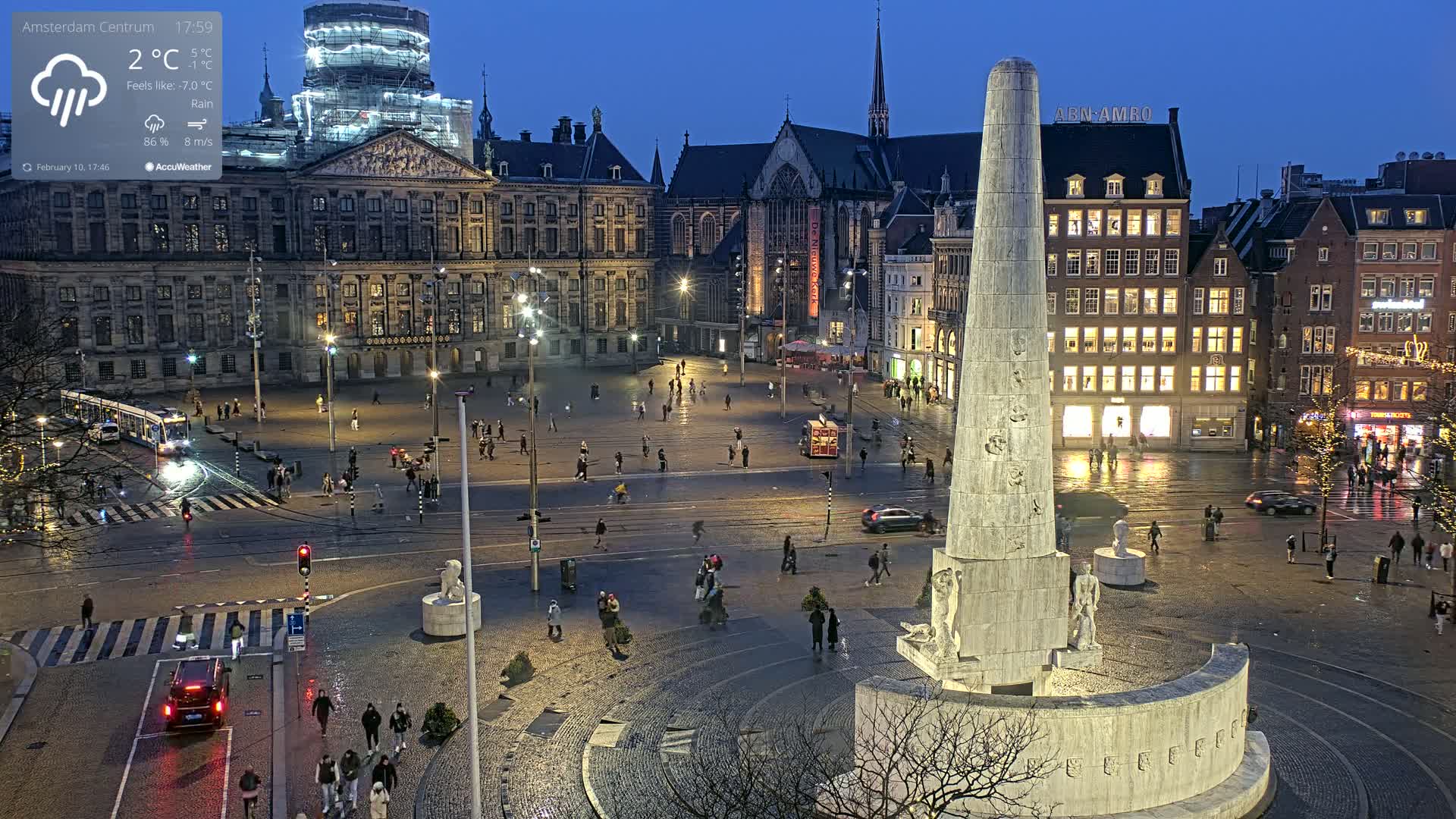 A nighttime view of a large, rain-slicked European city square with a tall monument at its center, surrounded by historic buildings and a scattering of pedestrians.
