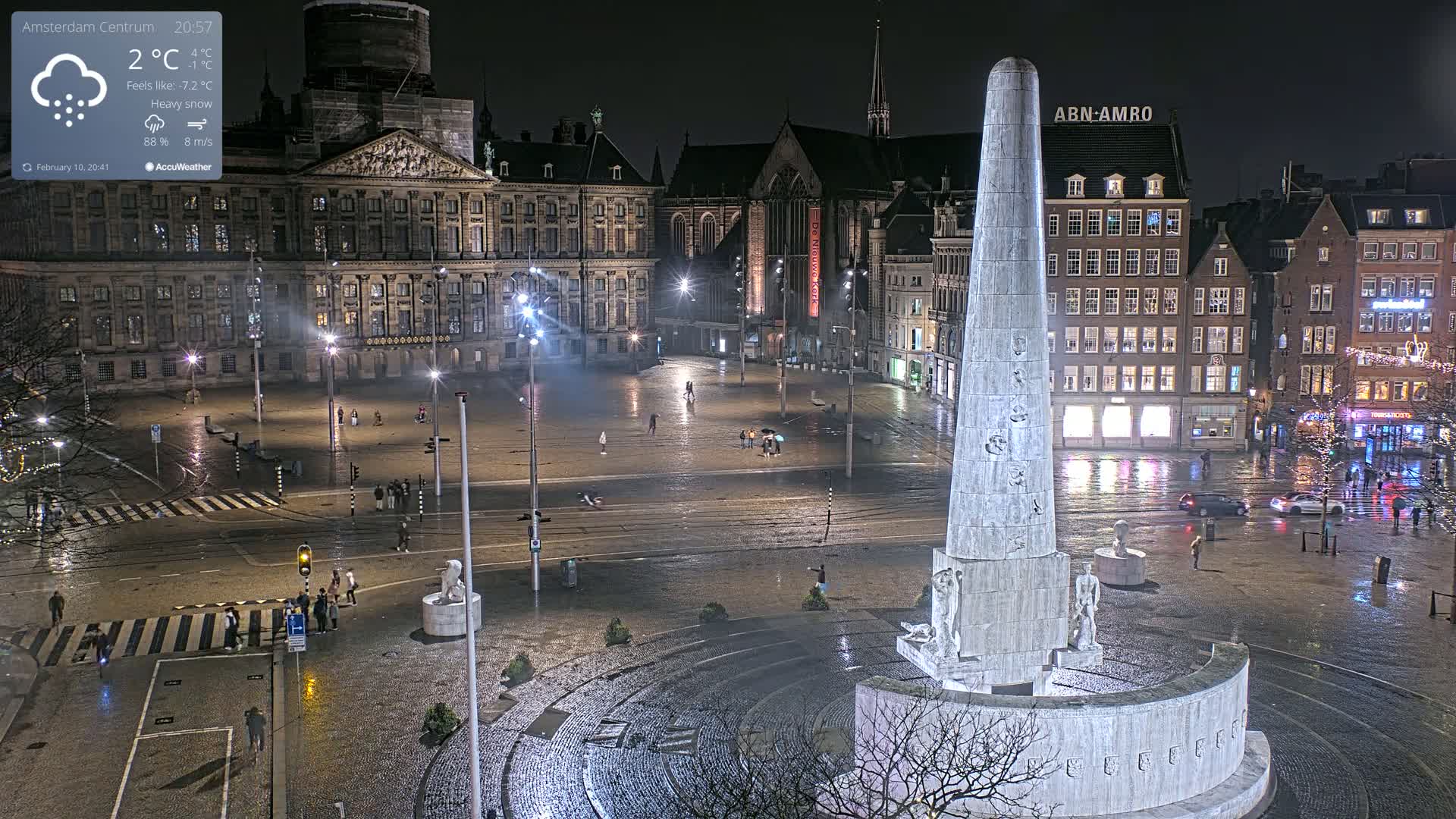 A nighttime view of a large, wet city square with a tall monument at its center, under heavy snow.