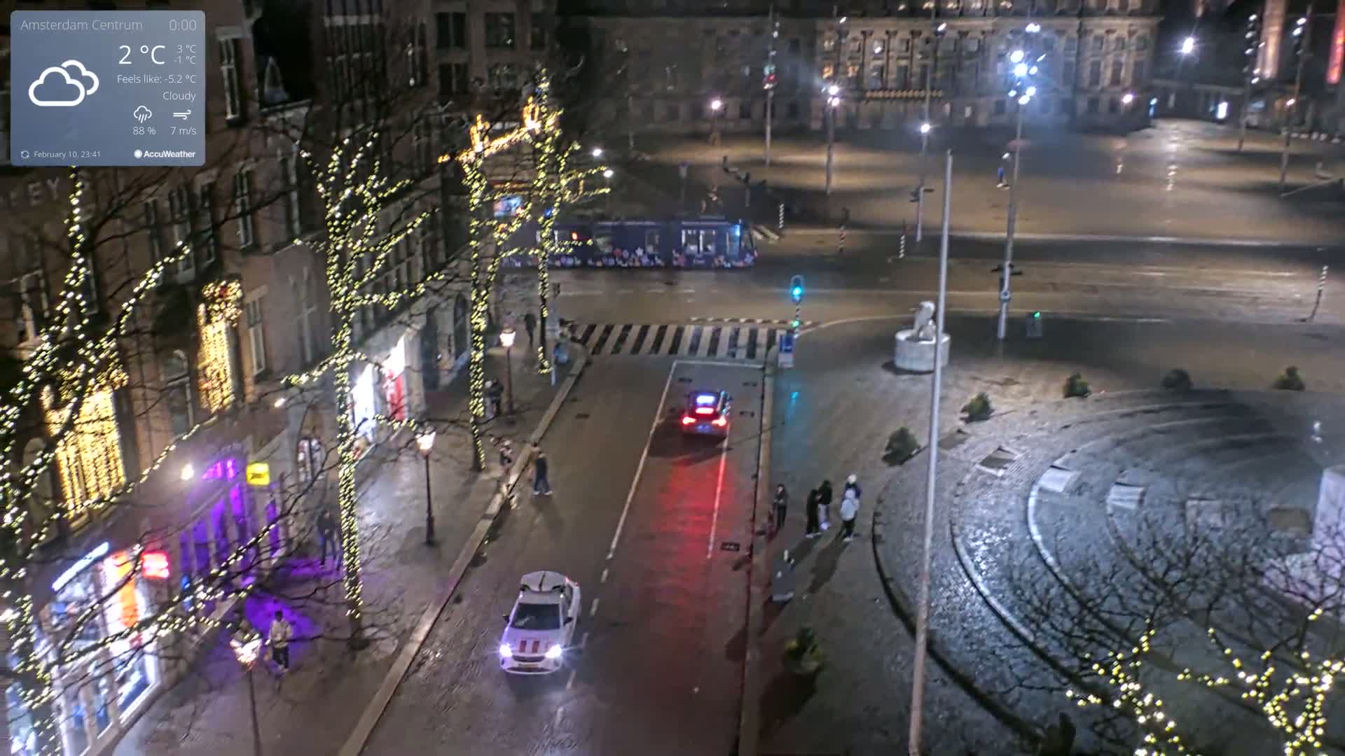 A nighttime, cloudy city scene shows a street with cars and pedestrians, buildings decorated with string lights, and a large open square.