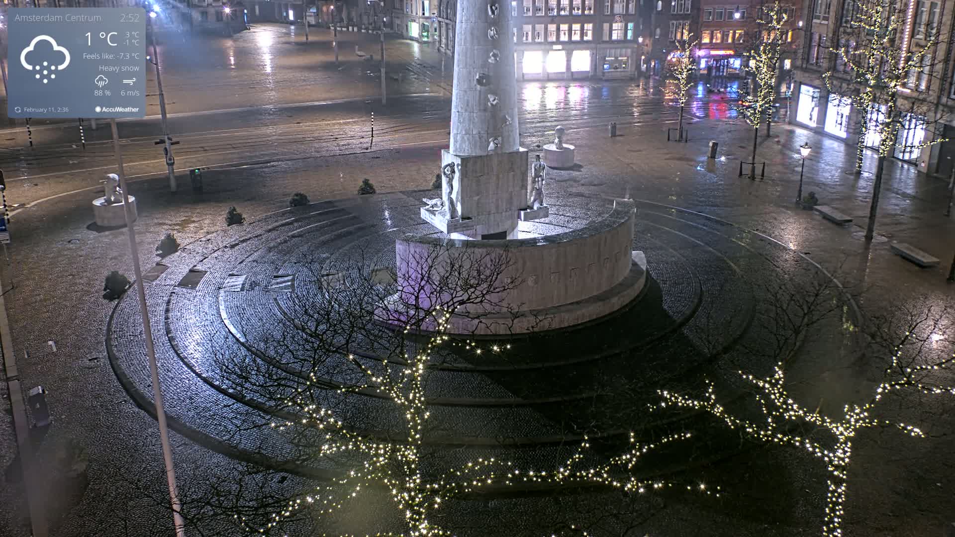 A monument in a city square is decorated with string lights under heavy snowfall at night.