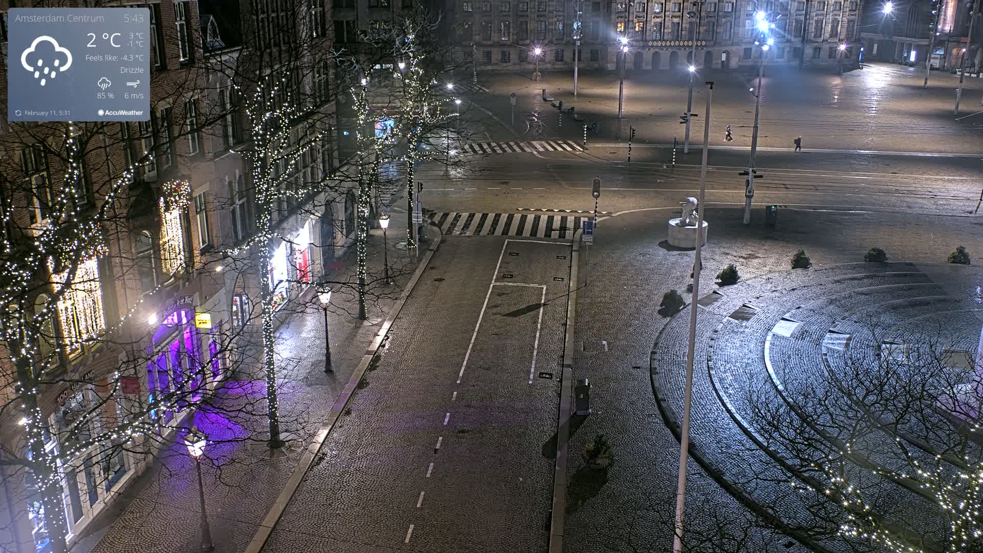 A nighttime view of a mostly empty cobblestone city square with string lights on trees lining the streets, under a drizzle.