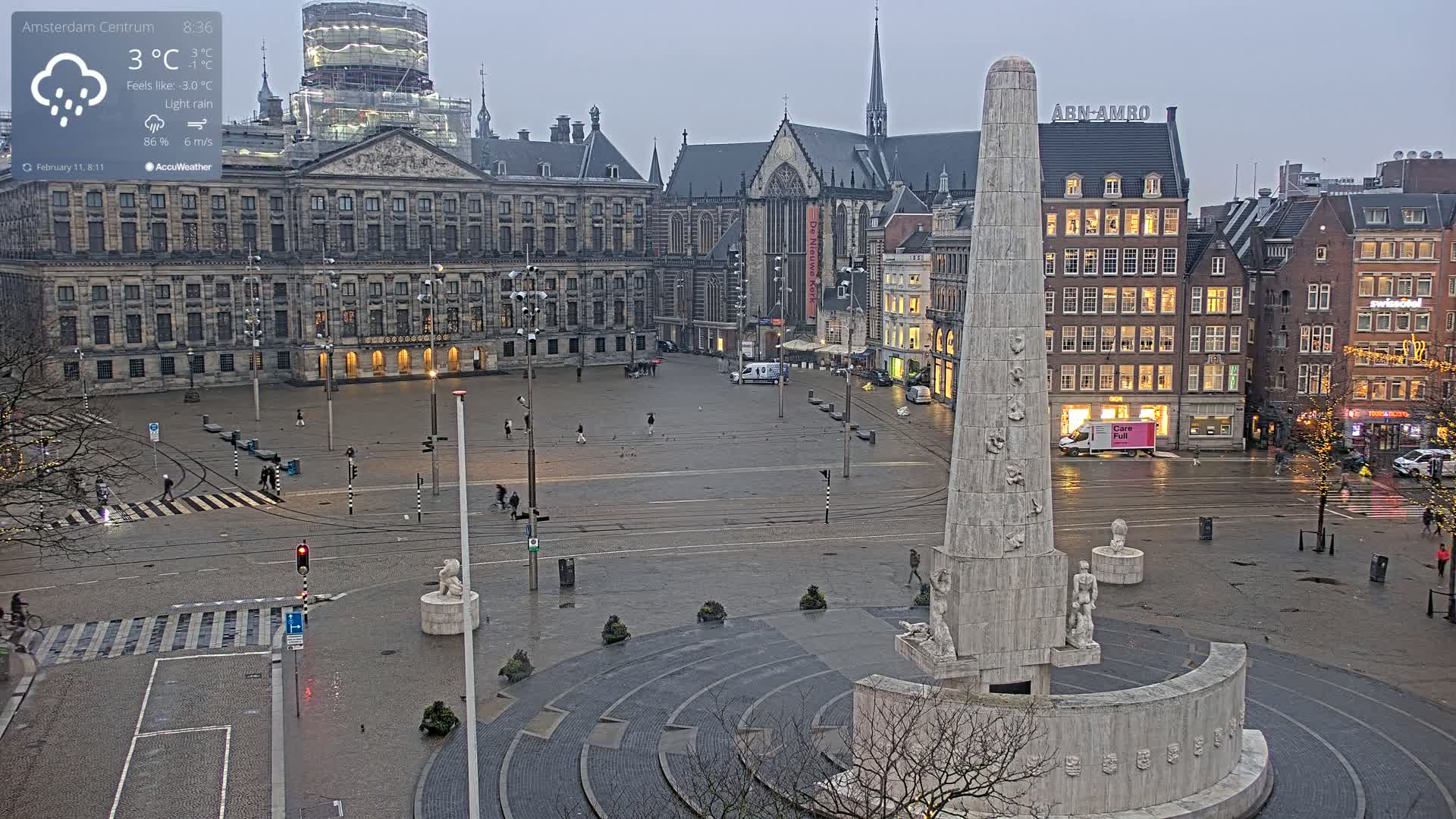 A large, wet city square is shown with a tall obelisk-like monument in the foreground, surrounded by historic buildings under a light rain.
