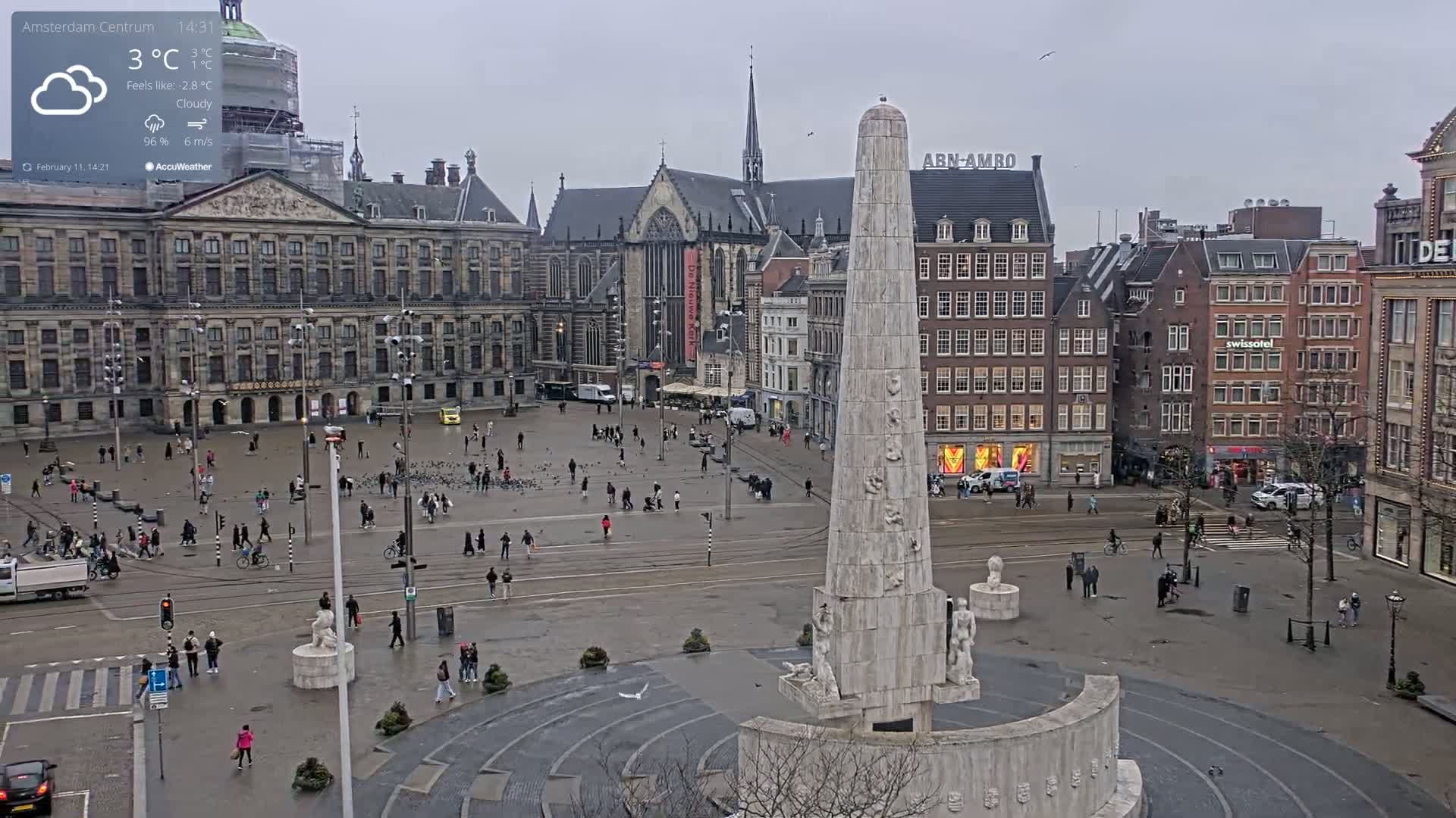 A cloudy, cool day in a large European city square, centered on a tall, light-colored monument surrounded by historical buildings and people walking around.