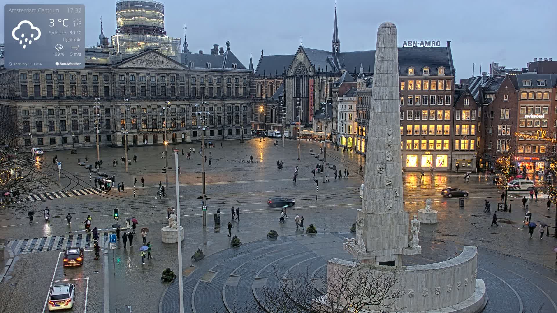 A rain-slicked city square, centered around a tall monument, is surrounded by buildings under a gloomy sky; people are sparsely scattered throughout the square.