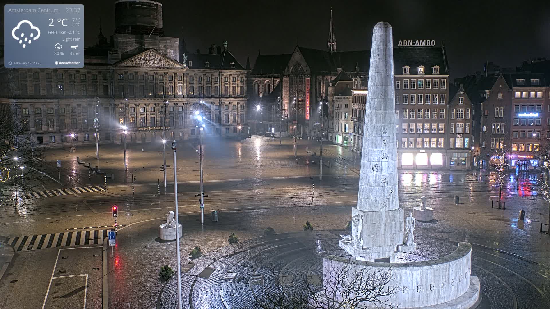 A nighttime view of a large city square, wet from light rain, features a tall monument in the foreground and a palace in the background.