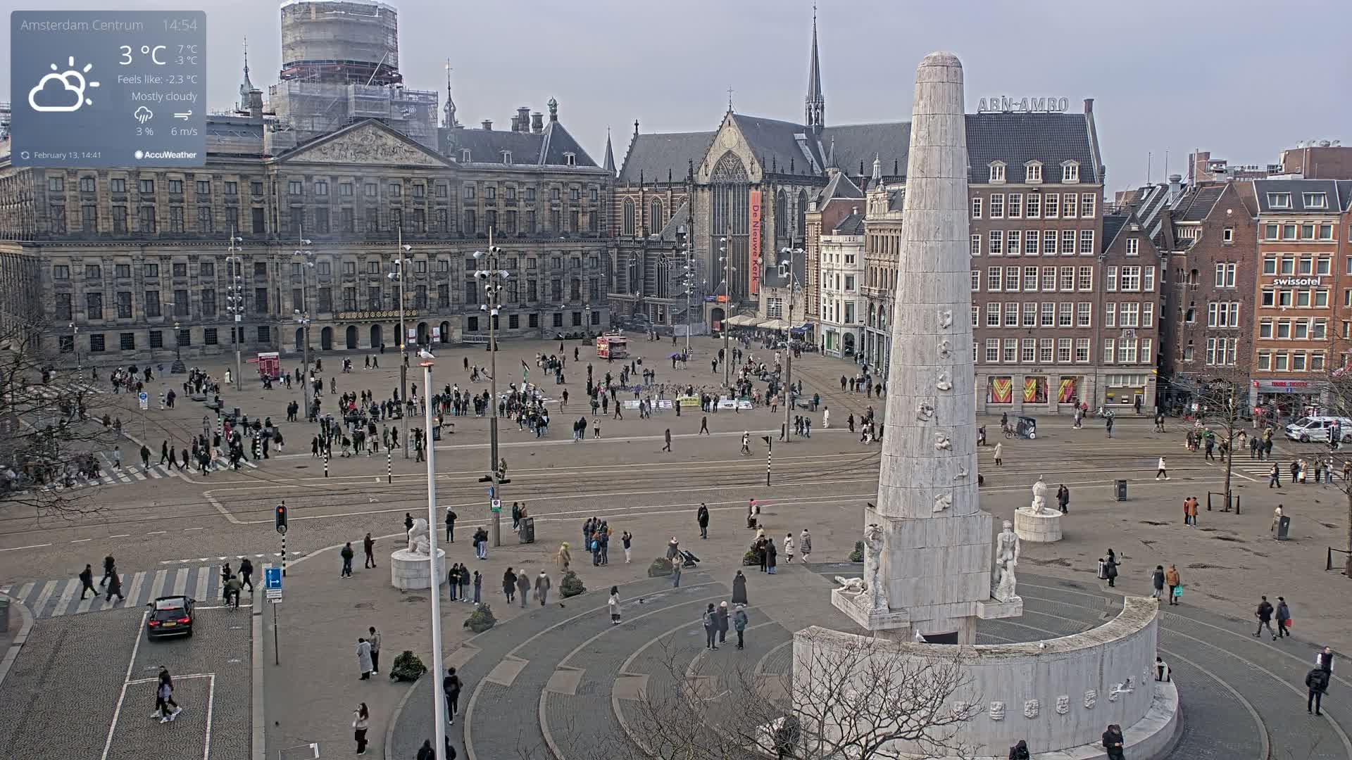 A mostly cloudy, cold day in a large European city square with a tall monument at the center, surrounded by historical buildings and people walking about.