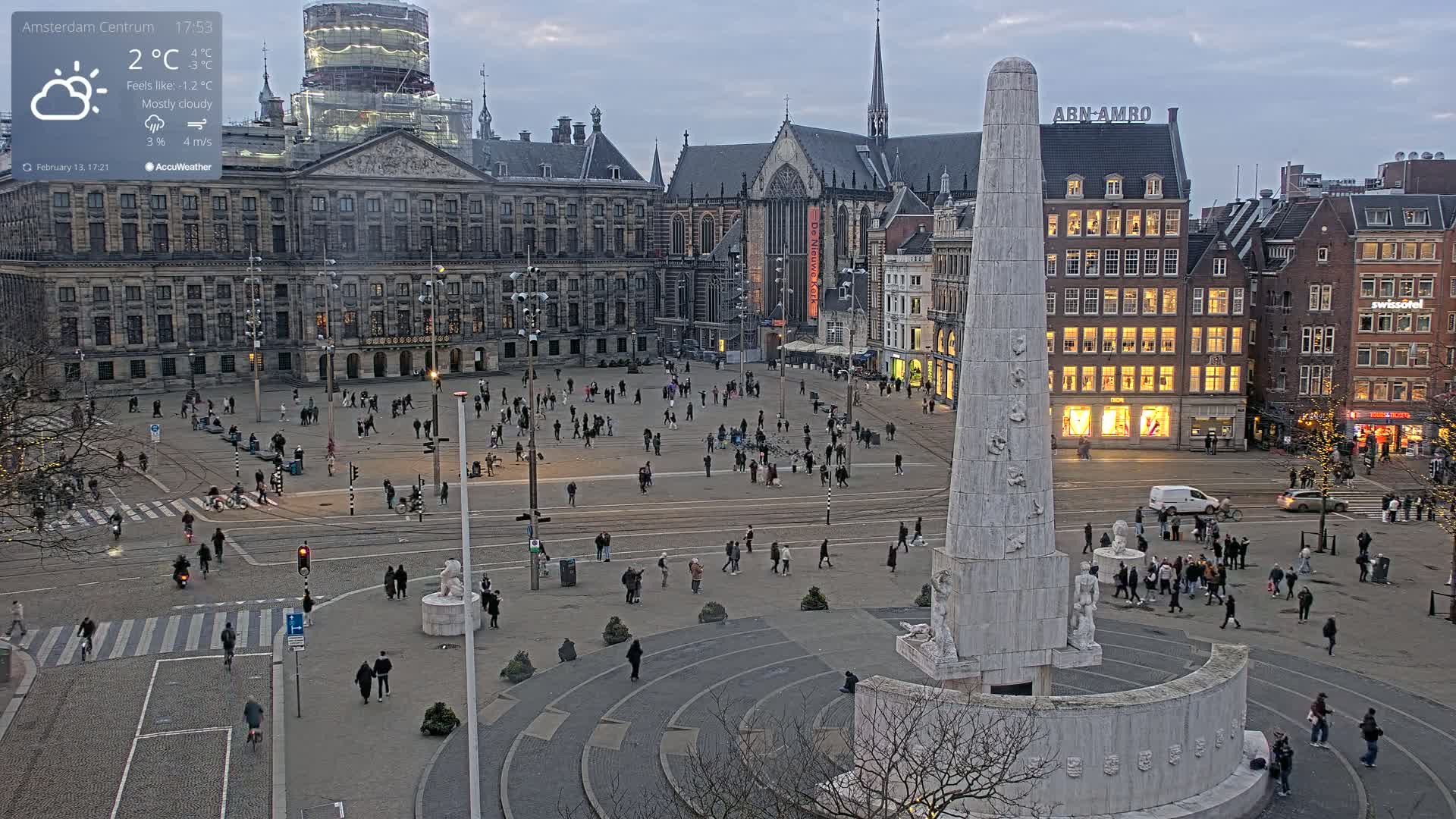 A mostly cloudy, cool evening scene shows a large city square with many people walking around a tall monument, buildings, and street traffic.