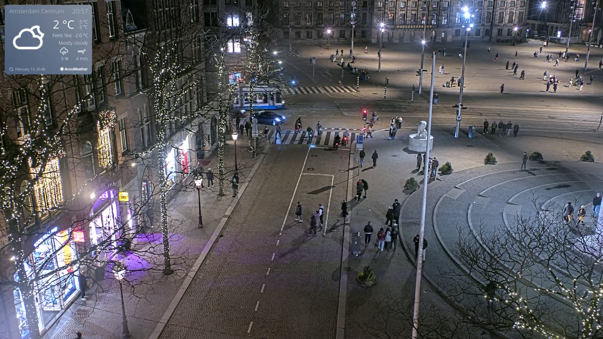 A mostly cloudy night scene shows a city square with pedestrians, streetlights, and Christmas lights strung along buildings and trees.