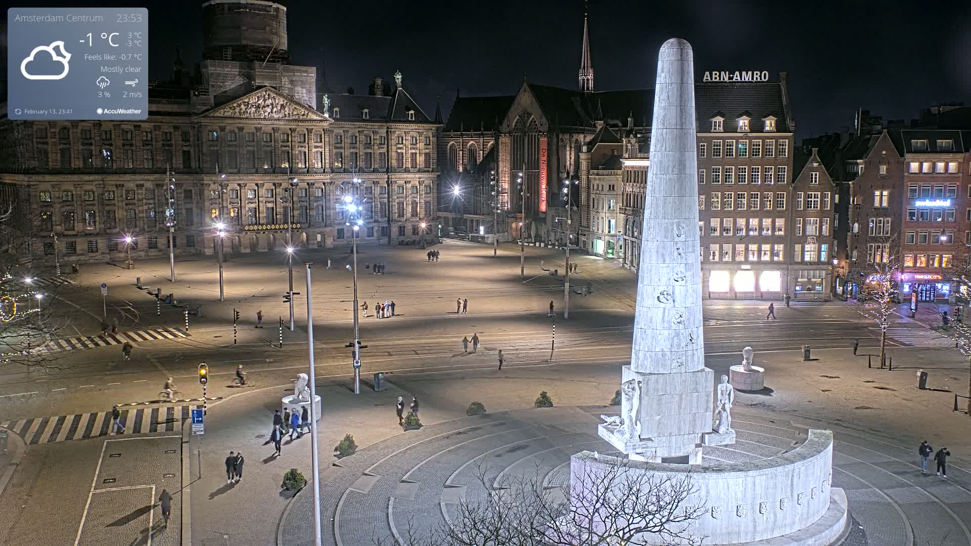 A nighttime, mostly clear view of a large, sparsely populated square in a city, centered on a tall, light-colored monument, with a large building and other structures in the background.