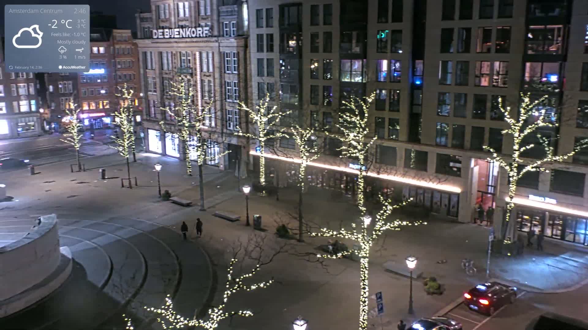 A mostly cloudy night scene shows a city square with bare trees adorned with white string lights, illuminated buildings, and a few pedestrians and cars.