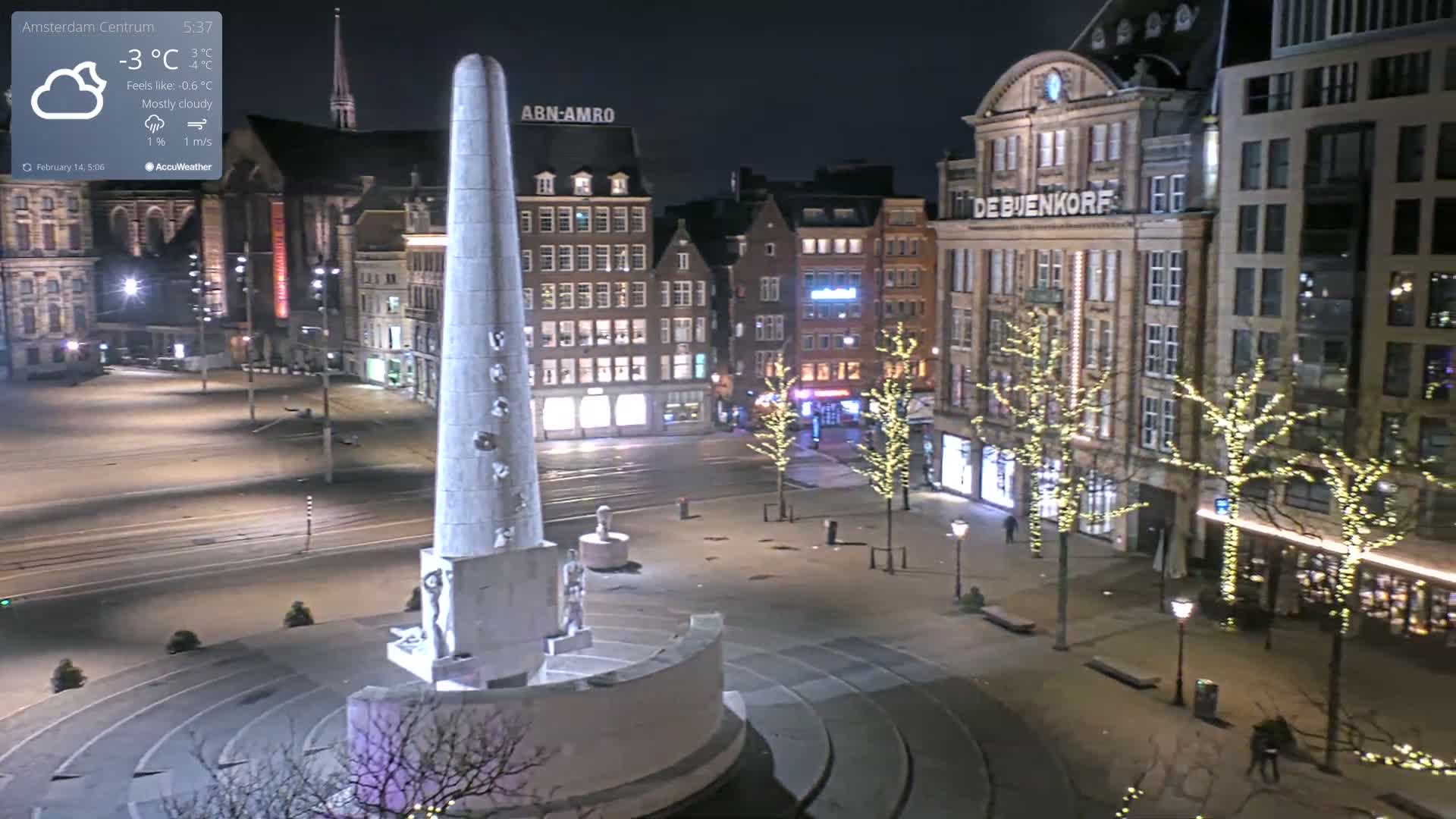 A mostly cloudy night scene shows a monument in a city square surrounded by buildings illuminated with lights, and some streetlights.