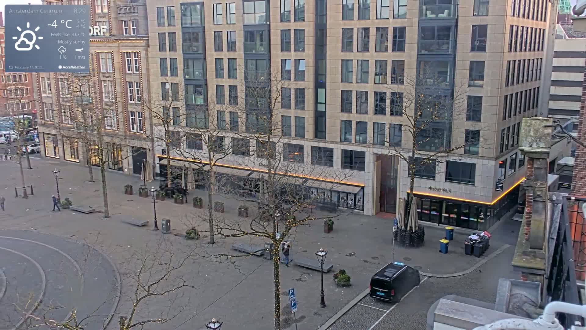 A mostly sunny, cold square in a city with bare trees, a few pedestrians, parked cars, and buildings with modern and older architecture.