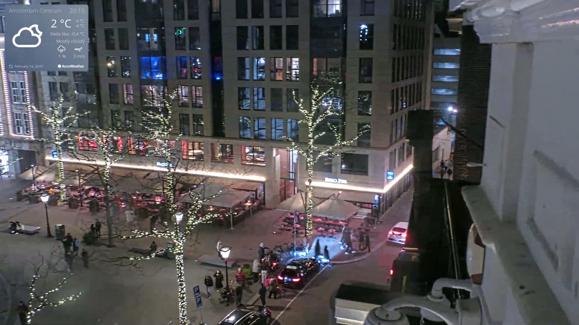 A mostly cloudy night scene shows a city square with several restaurants and trees decorated with string lights, people walking around, and cars parked along the street.