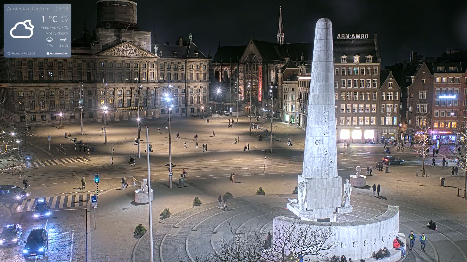 A mostly cloudy night scene shows a large, illuminated city square with a tall monument at its center, surrounded by buildings and a few pedestrians.