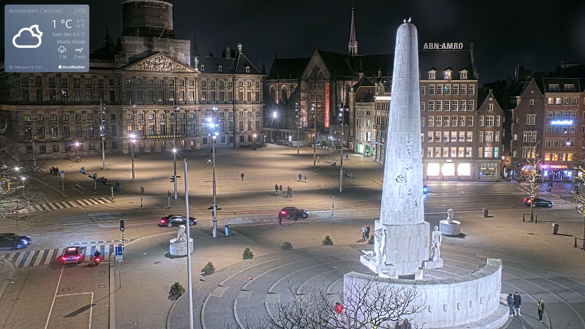 A mostly cloudy night scene shows a large, empty square with a tall monument in the foreground, surrounded by illuminated buildings.