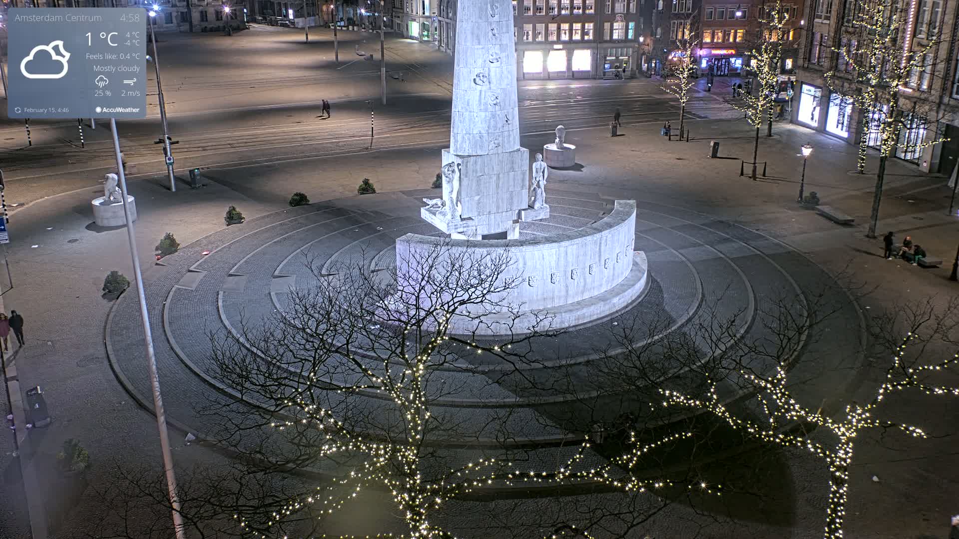 A mostly cloudy nighttime scene shows a large, circular monument with figures at its base, surrounded by bare trees with string lights and a paved plaza.