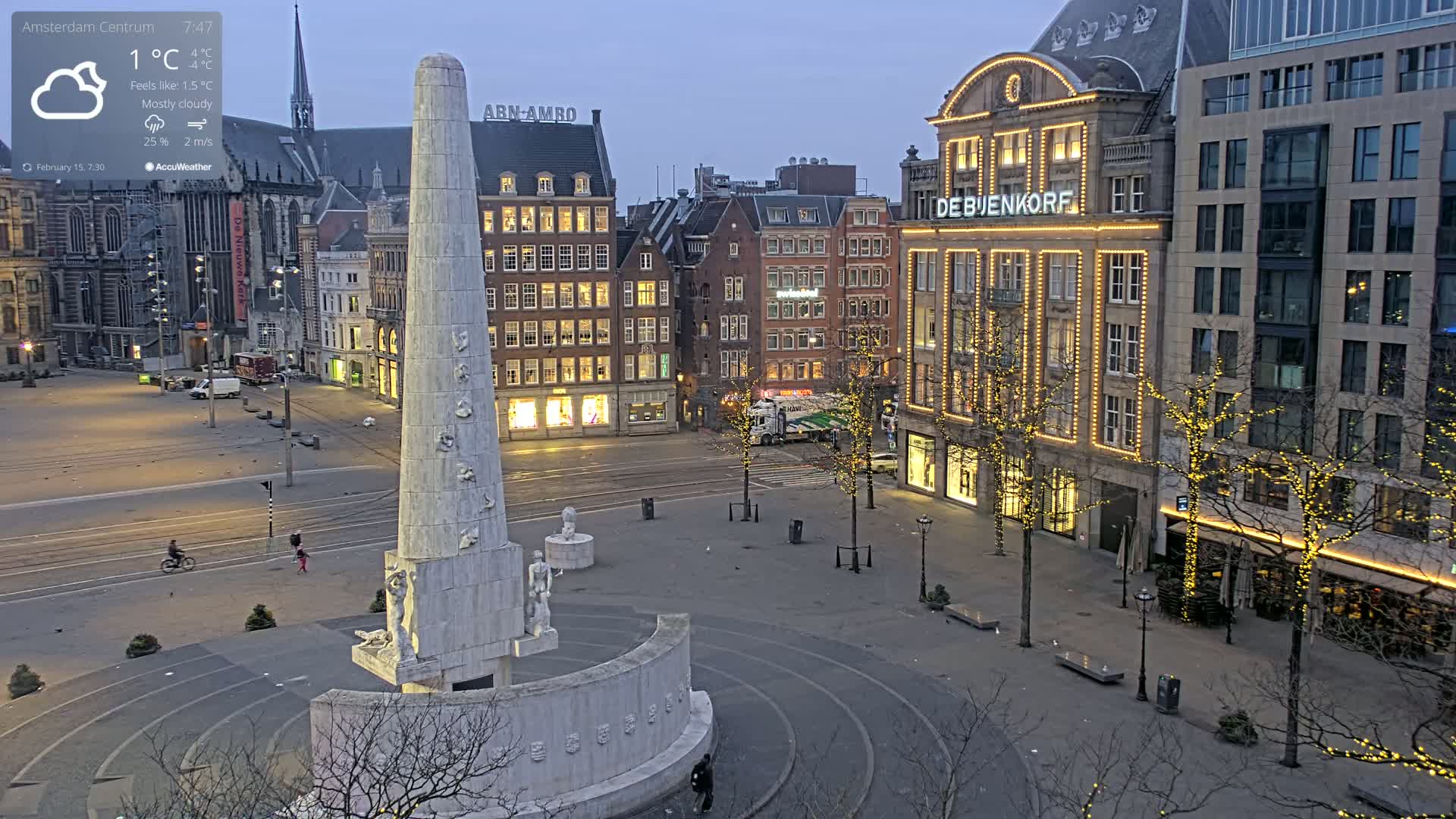 A mostly cloudy, cool evening scene shows a large city square with a tall monument at its center, surrounded by buildings with illuminated storefronts and trees strung with lights.