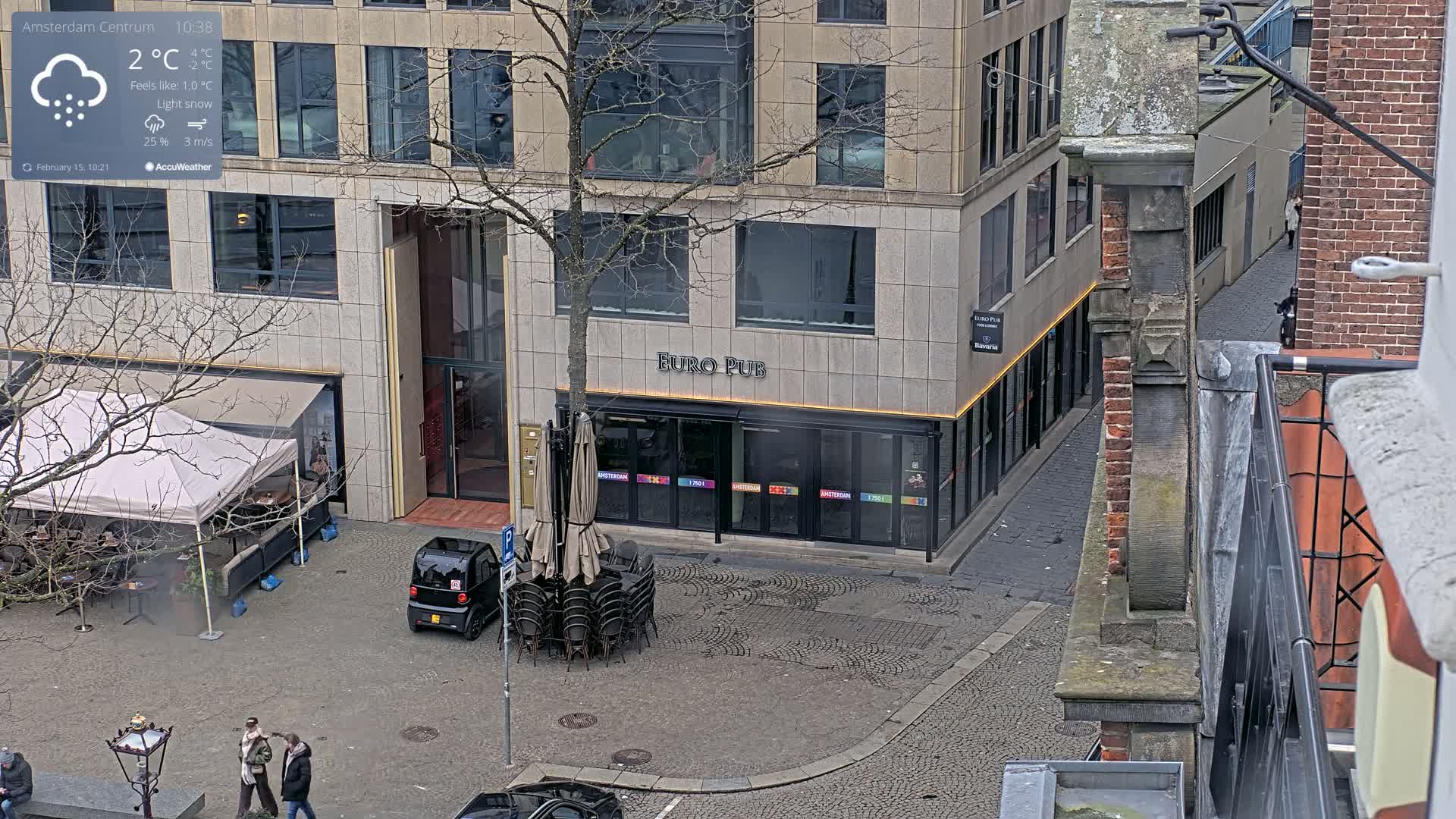 A city street scene with light snow falling shows a Euro Pub with closed patio umbrellas and a small car parked nearby, next to a modern building and an older brick building.